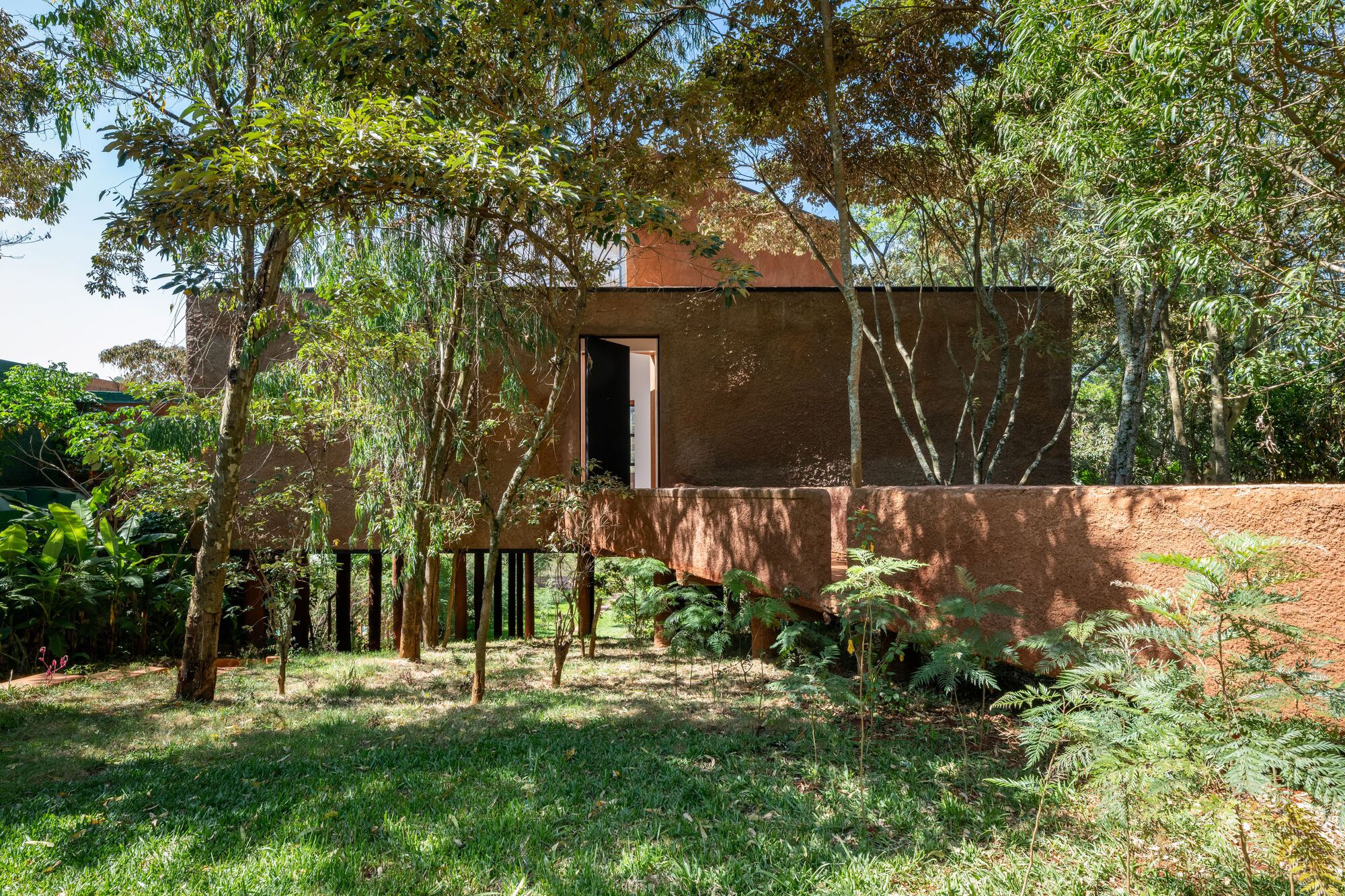 Ground-level view of the brick volume on black pilotis flanked by young trees and dappled sunlight