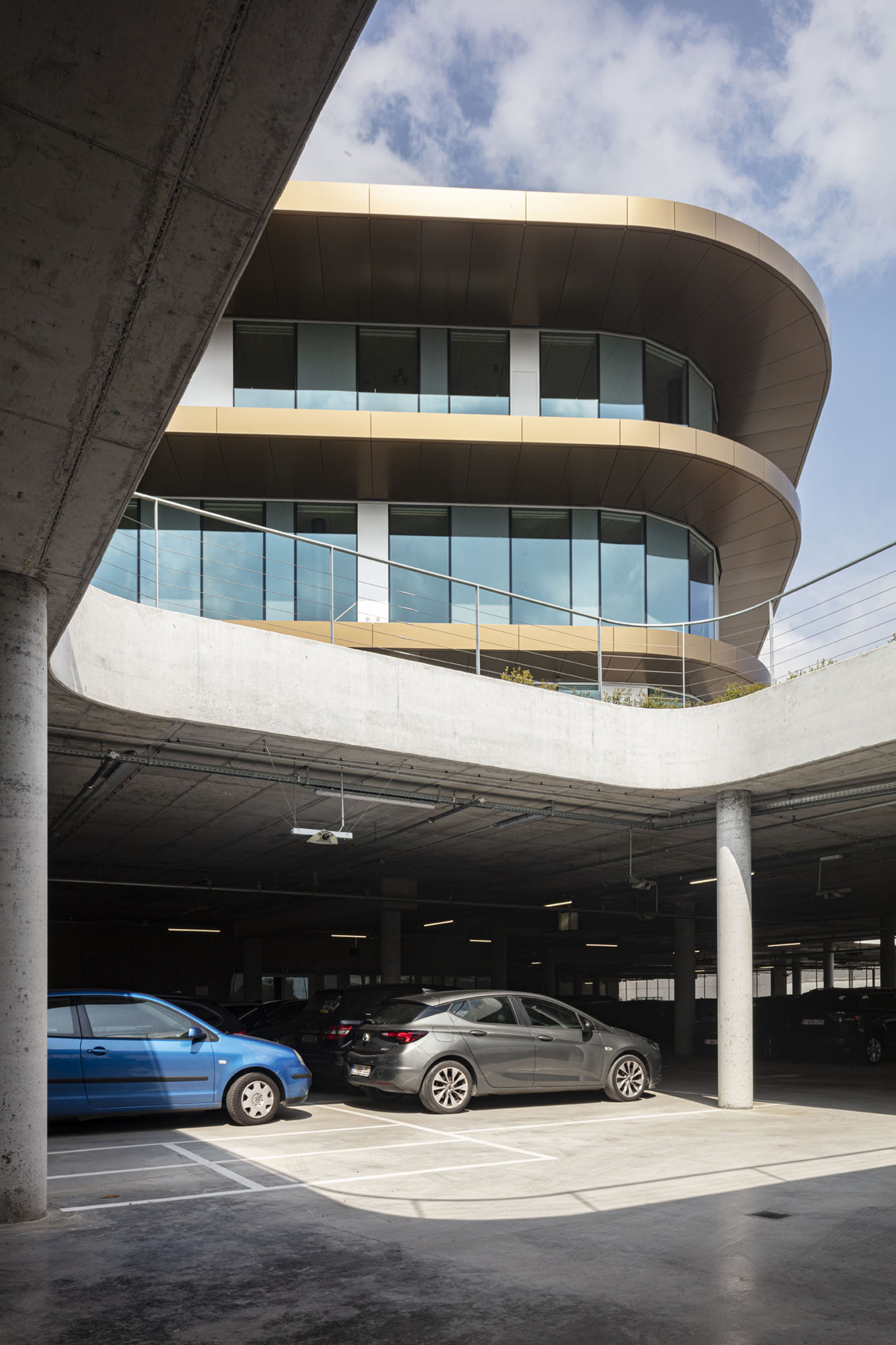 Rounded glass facade rising above an exposed concrete parking structure with parked cars
