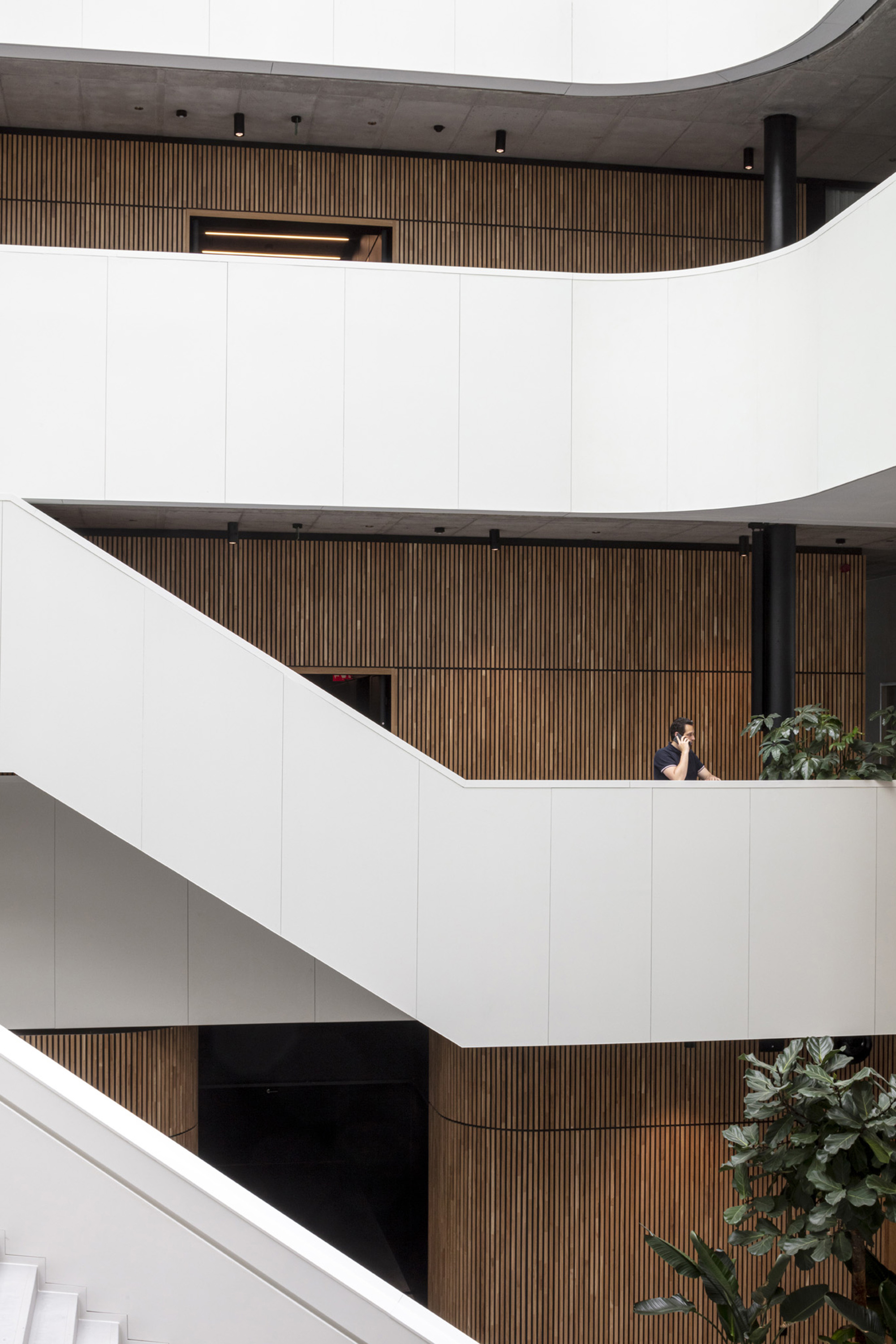 Stacked white balconies with timber slat cladding and a person seated at mid-level next to potted plants