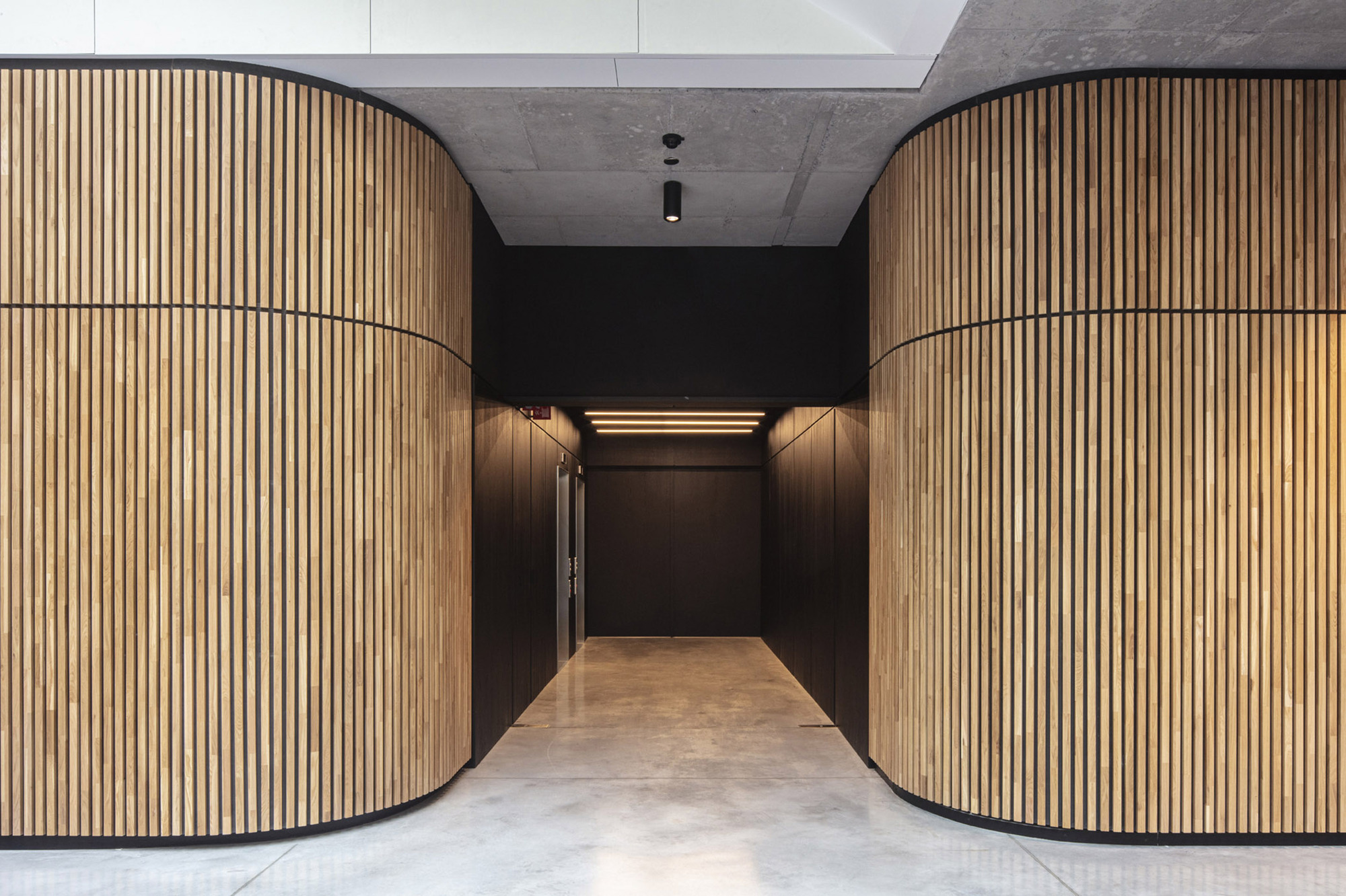 Corridor framed by curved timber slat walls leading to a dark-paneled elevator lobby under concrete ceiling