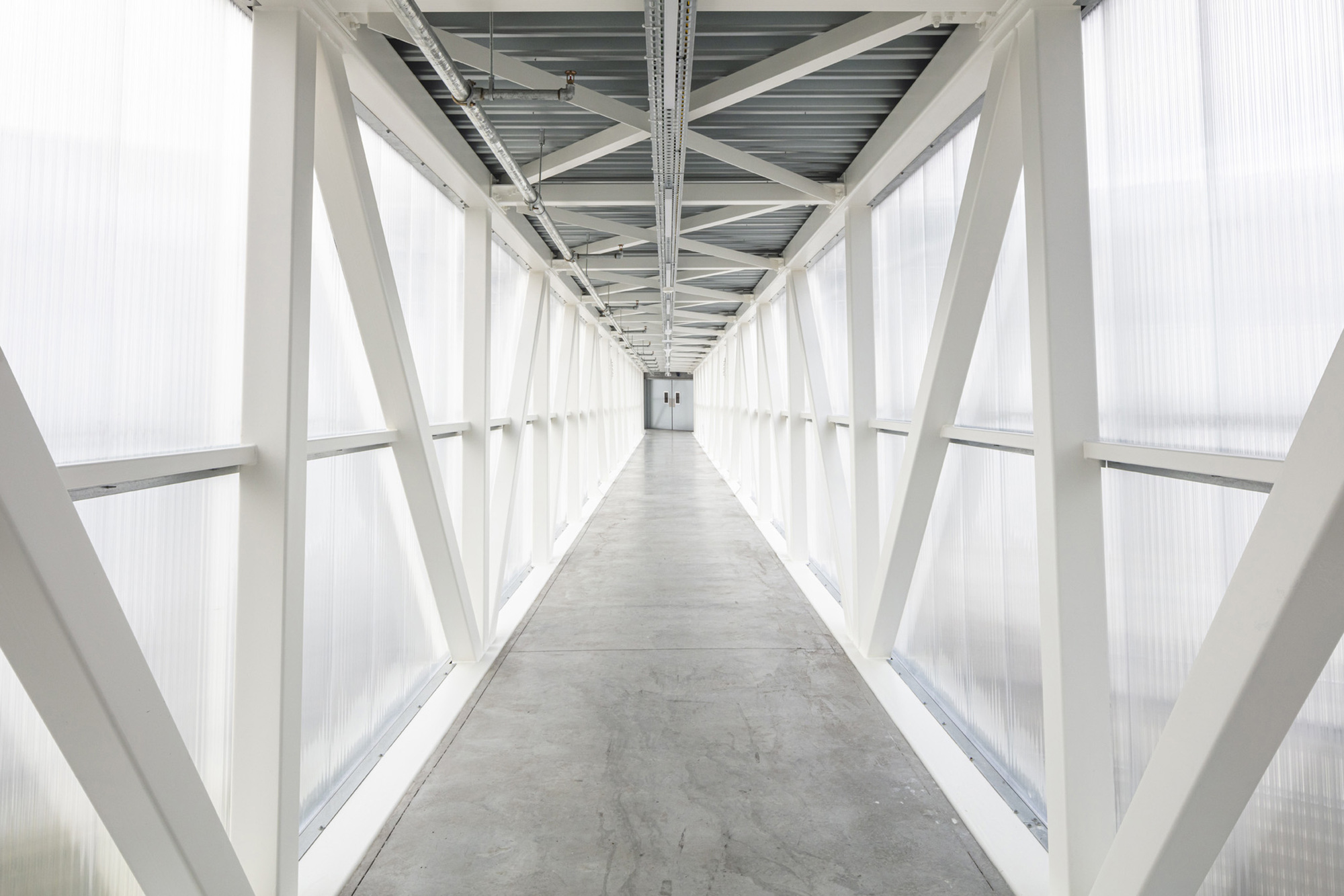 Enclosed corridor with white diagonal steel bracing and translucent polycarbonate cladding under an overcast sky