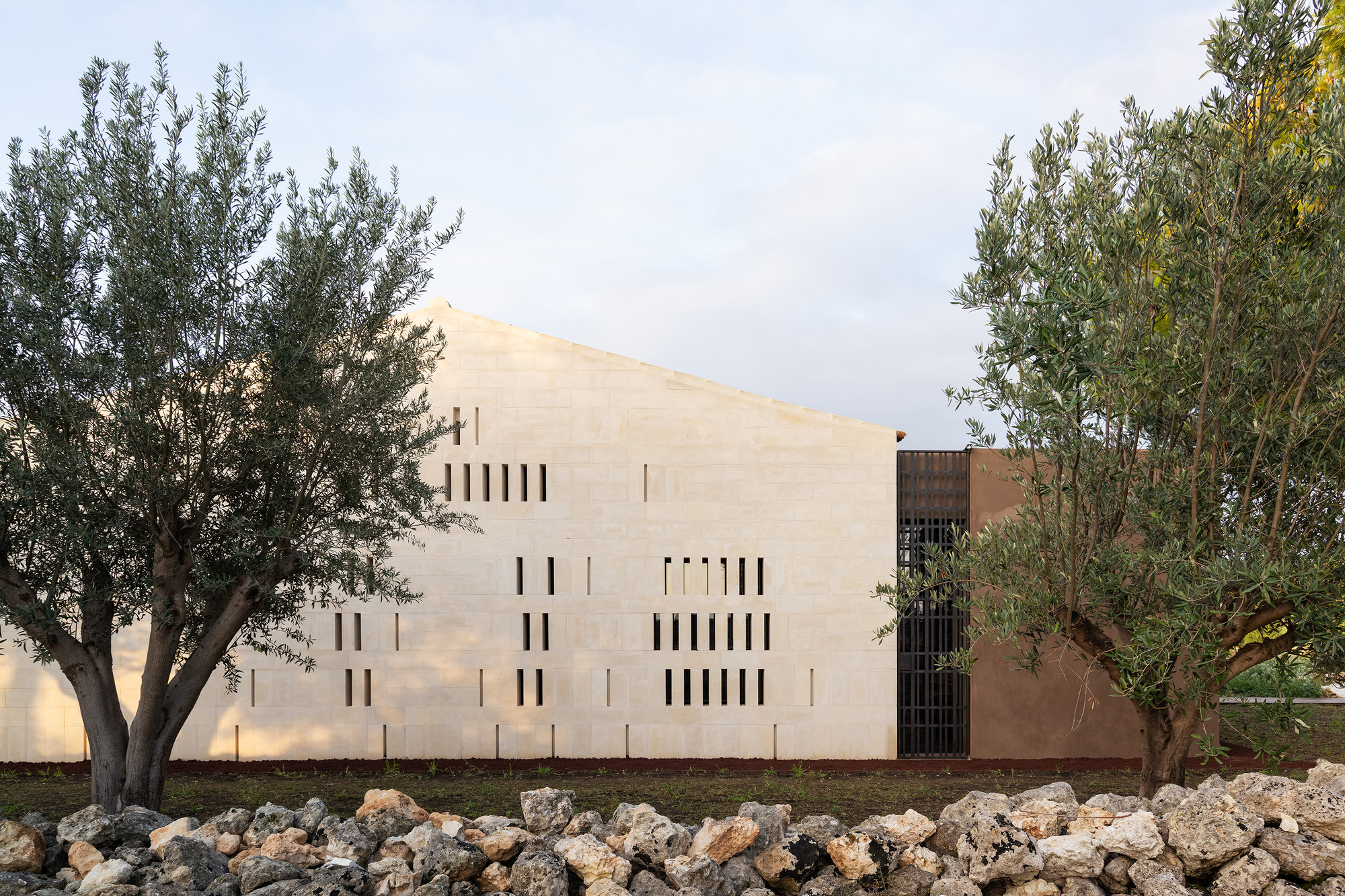 Pale rendered facade with vertical slot windows framed by olive trees and rocky ground