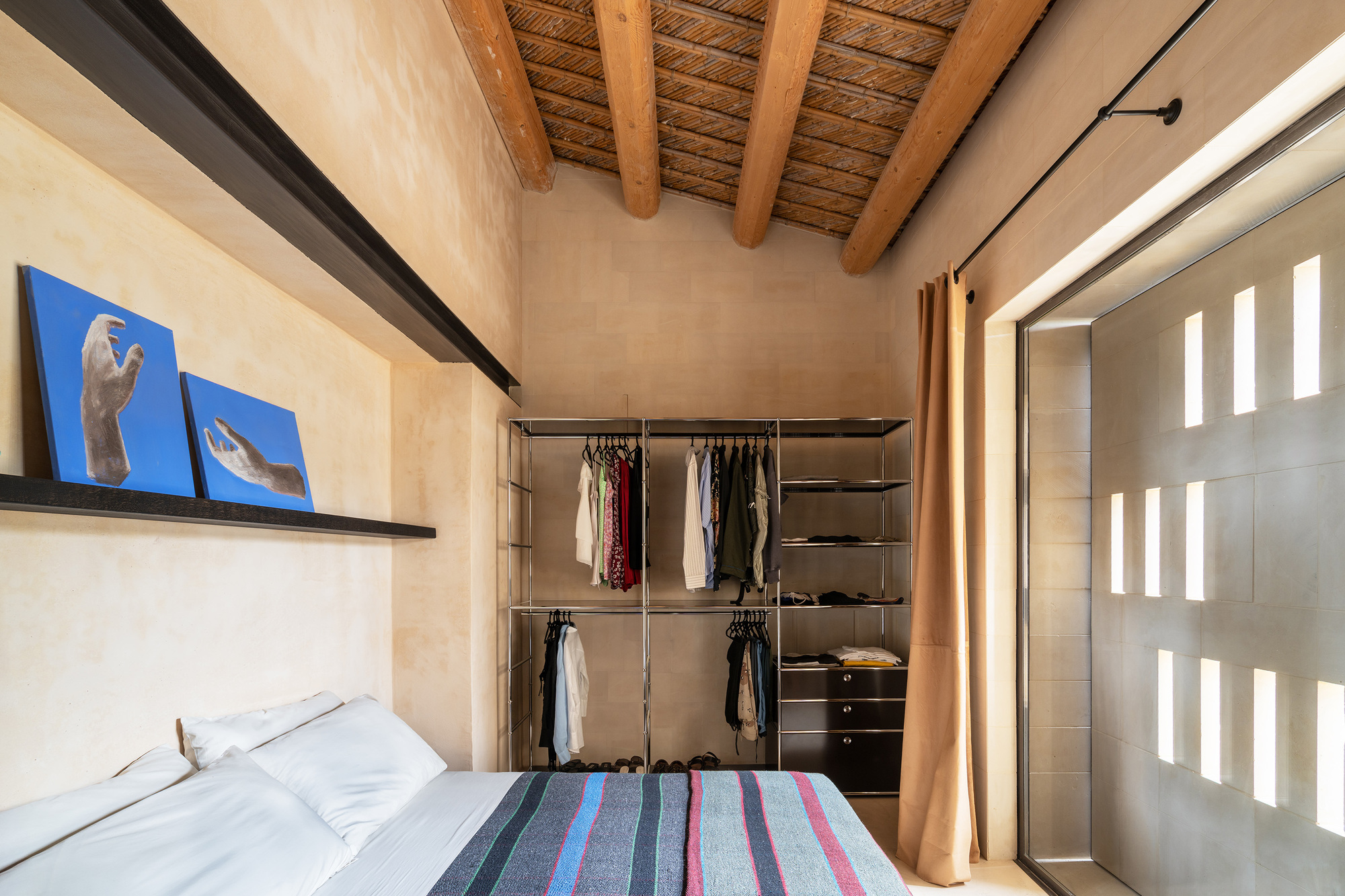 Bedroom with exposed timber ceiling beams and open wardrobe screened by curtain beside tall window slits