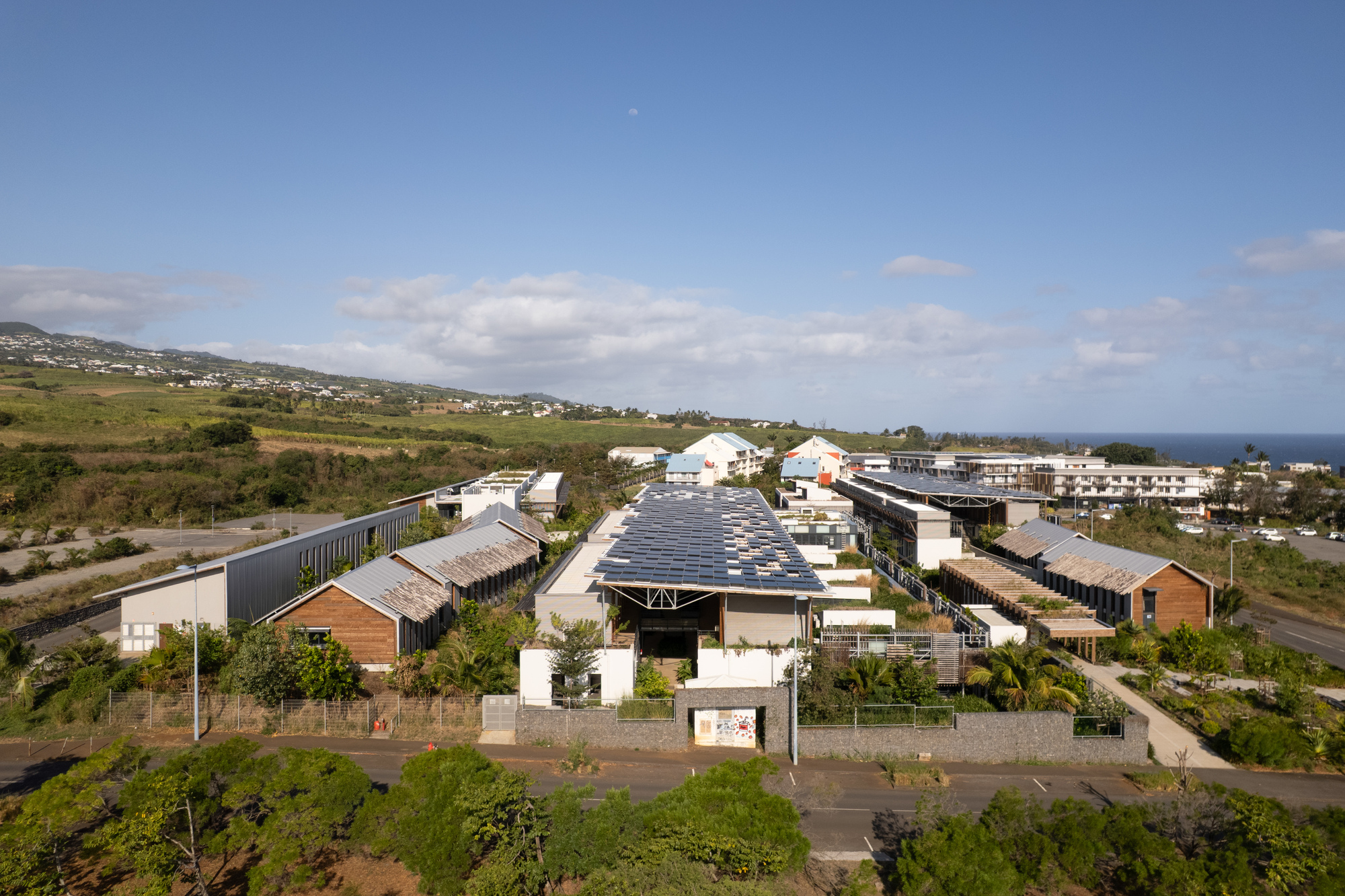 Distant view of the complex with multiple gabled roofs and photovoltaic panels against a hillside and ocean
