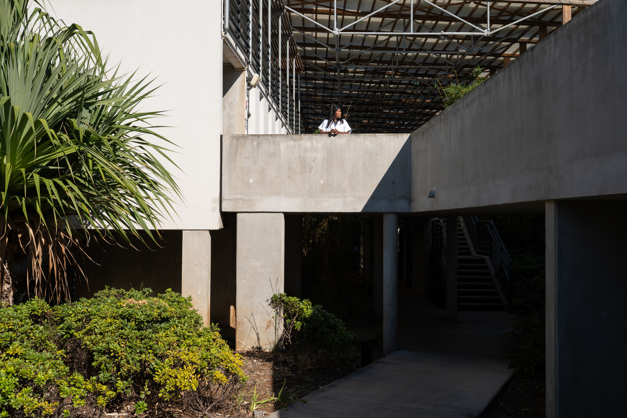 Concrete terrace with overhead trellis and two figures standing on elevated balcony above shadowed passage