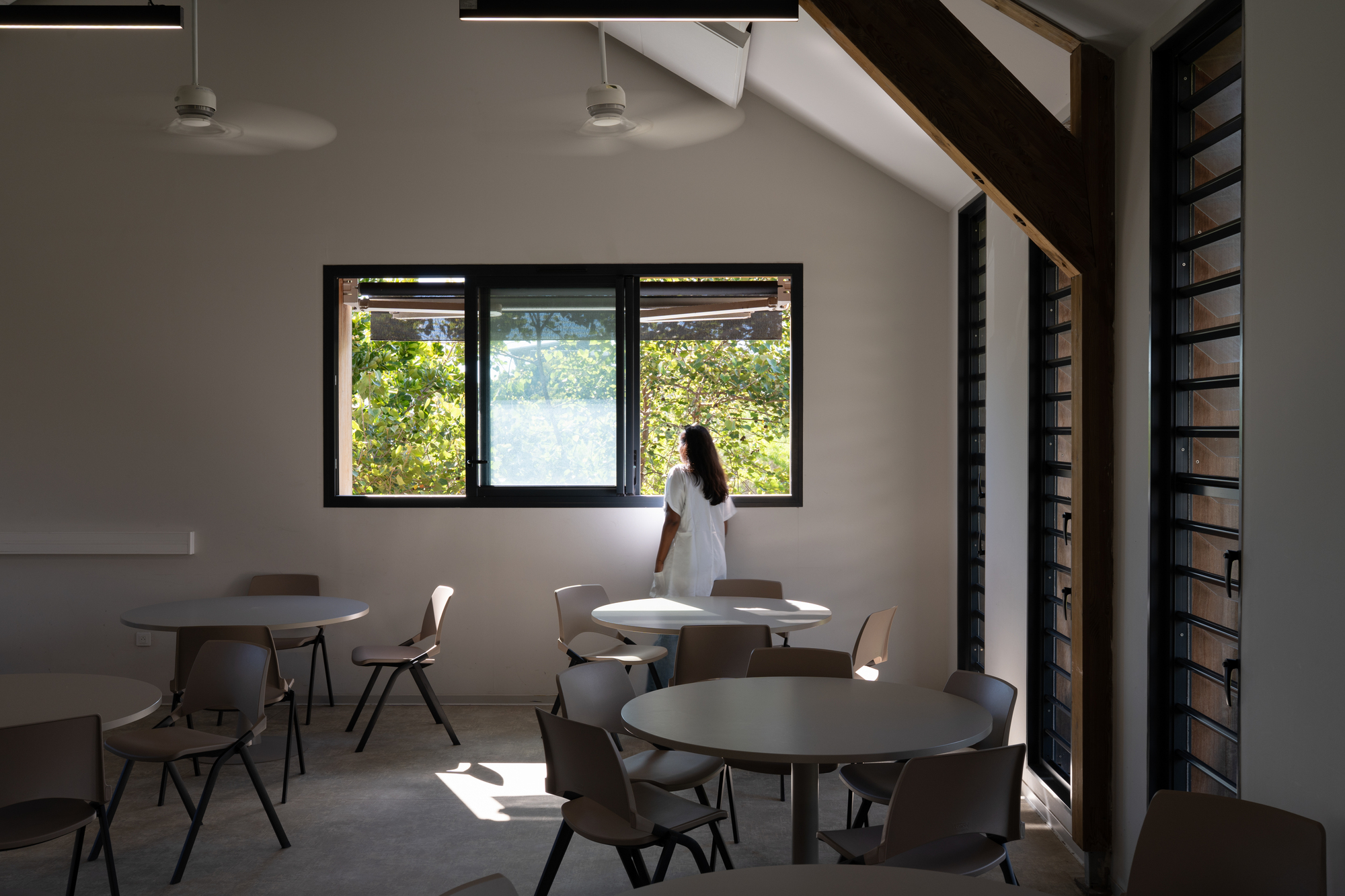 Dining area with round tables and a person standing by windows framing green foliage outside