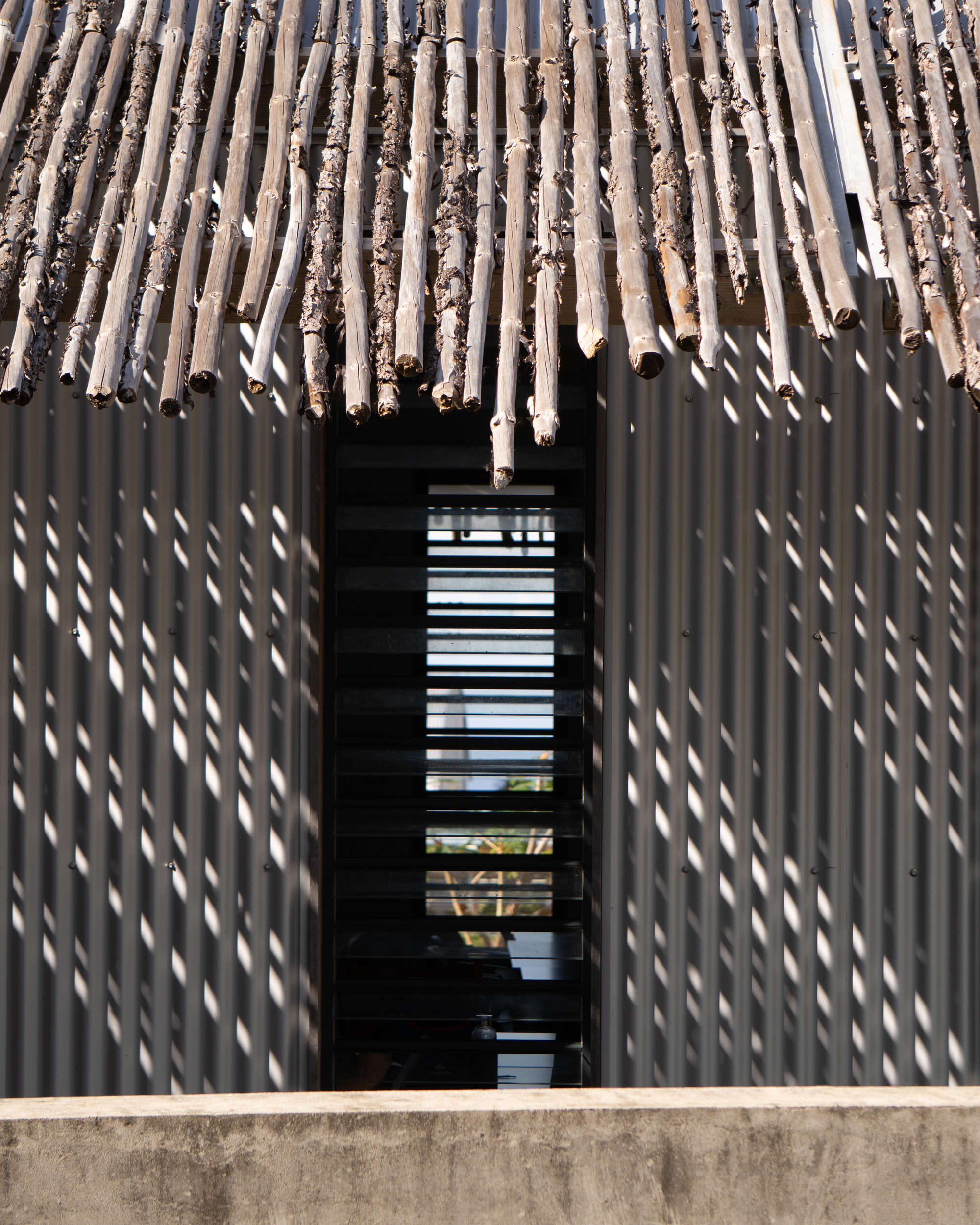 Facade of vertical timber slat screen with metal louvered window opening casting shadows in sunlight
