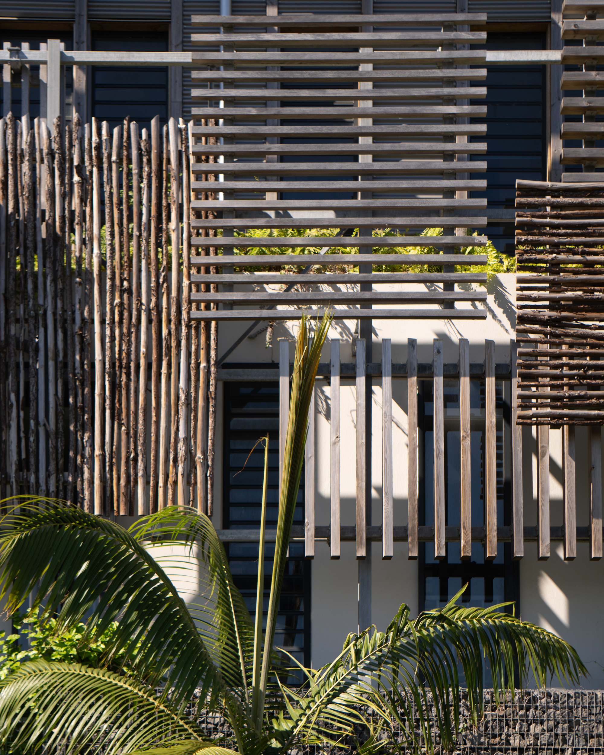 Layered screening system combining weathered timber branches and horizontal metal slats with palm fronds below