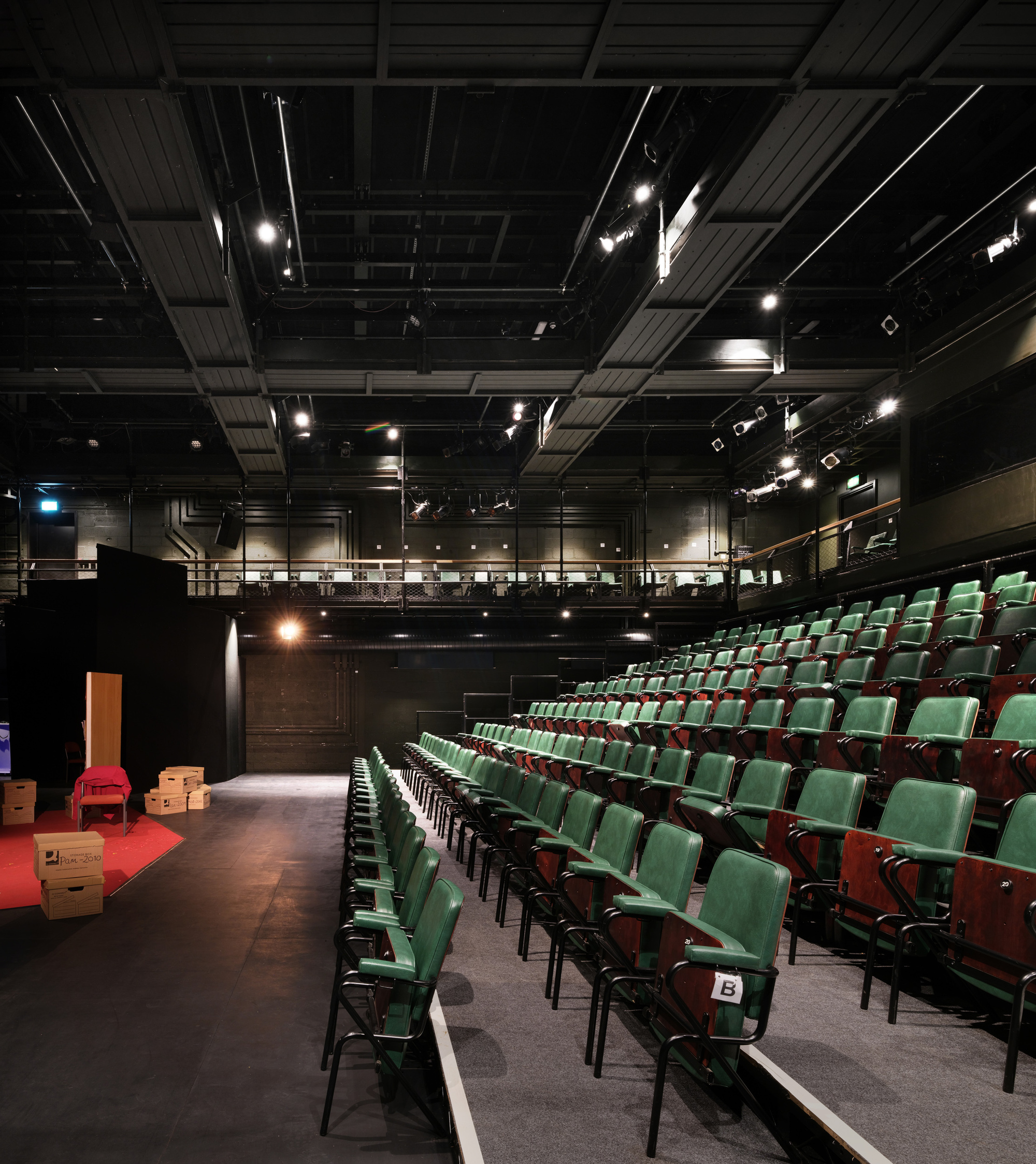 Theater auditorium with raked seating, exposed black ceiling with stage lights, and view toward the stage