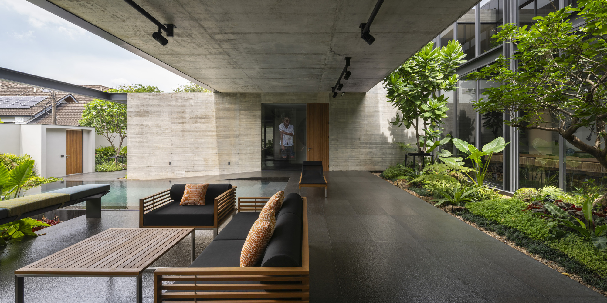 Covered outdoor lounge beneath exposed concrete soffit with timber furniture adjacent to lap pool and planted beds