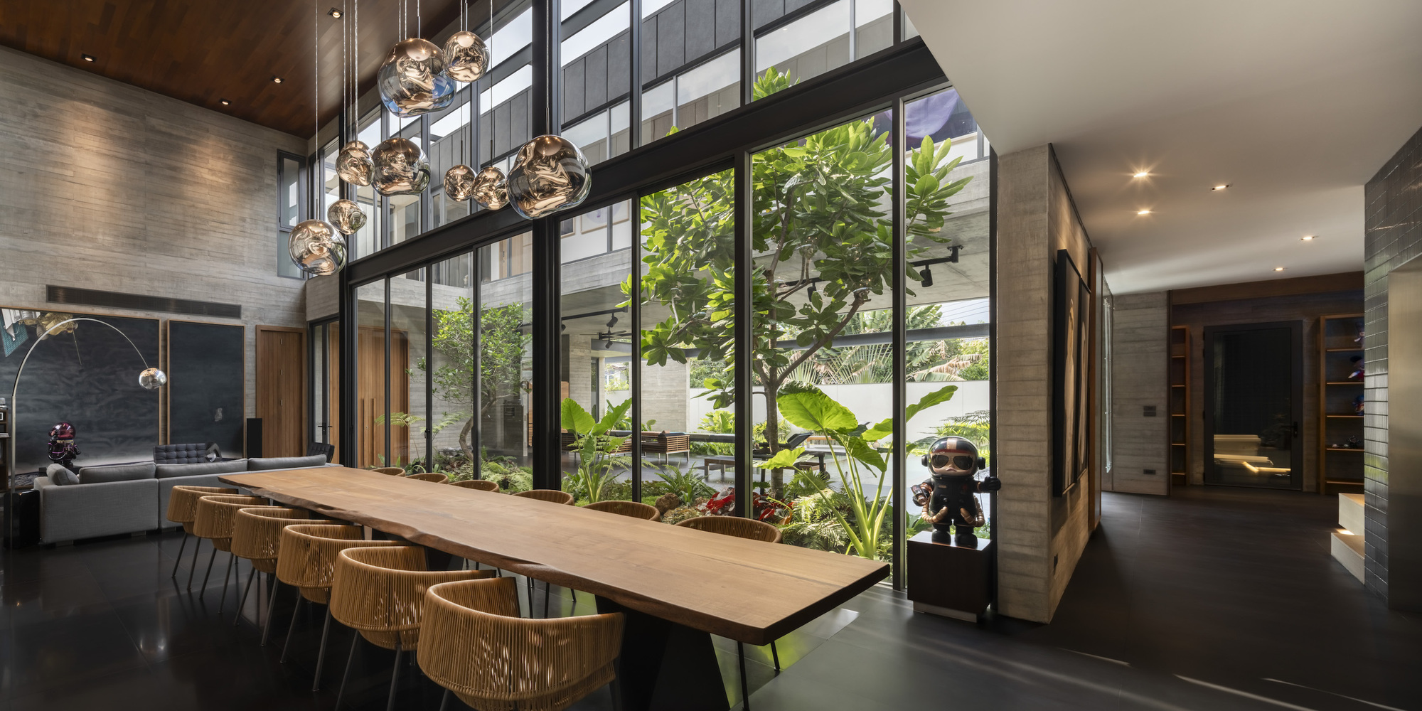 Dining space with long timber table beneath clustered pendant lights adjacent to a planted atrium