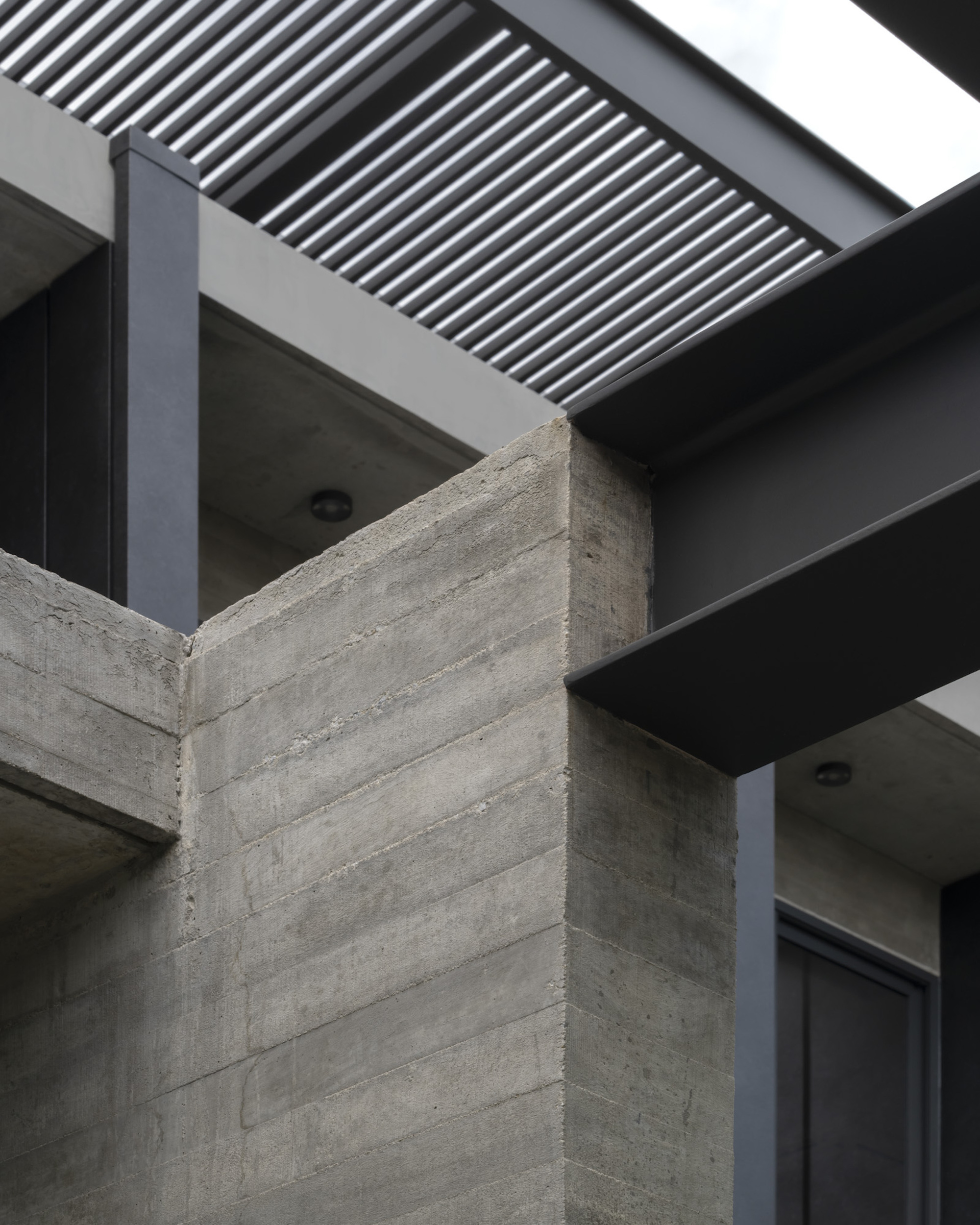 Board-formed concrete corner column meeting dark metal beams and overhead louver screen