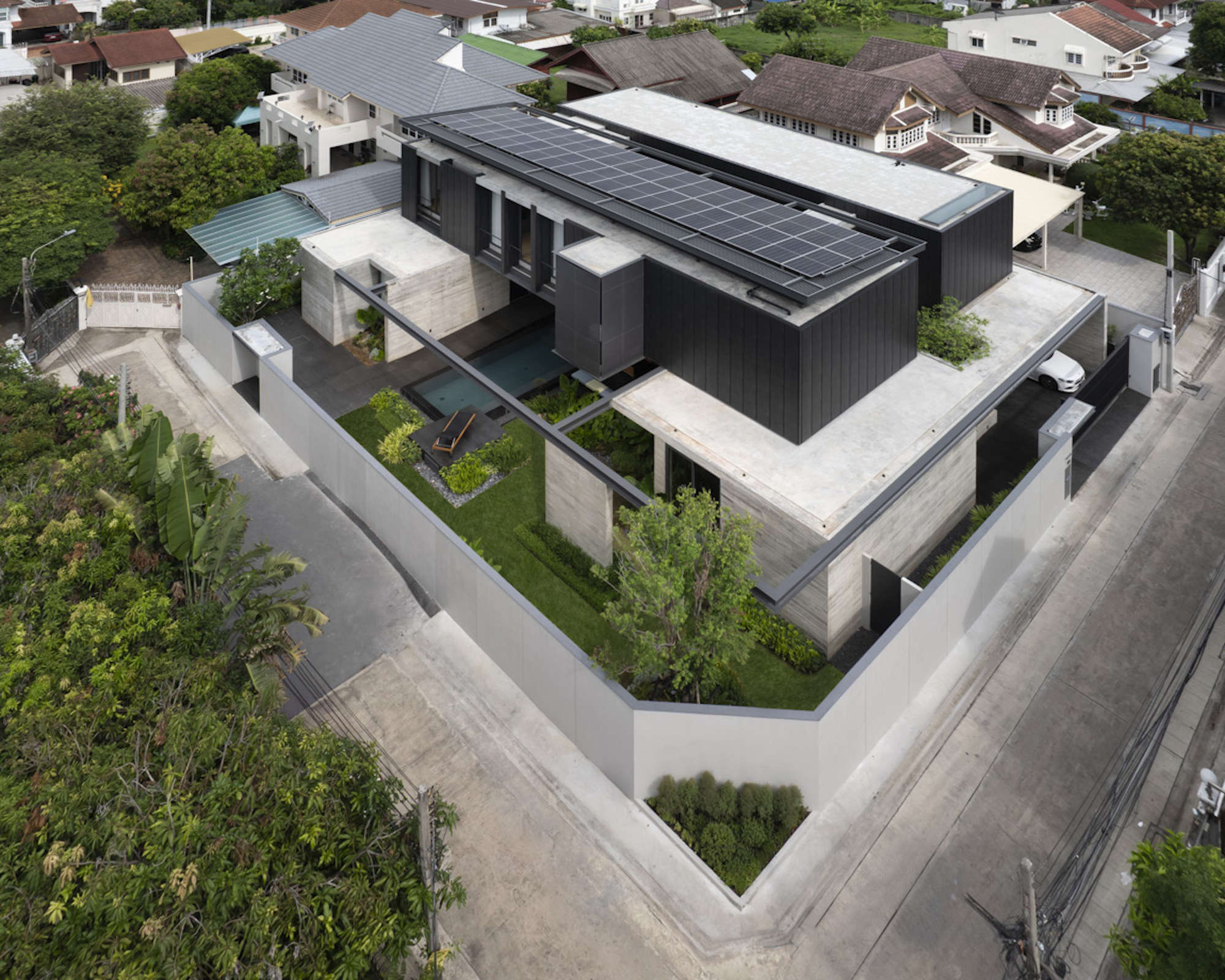 Aerial view of dark timber volume with solar panels surrounded by concrete terraces and landscaped courtyards
