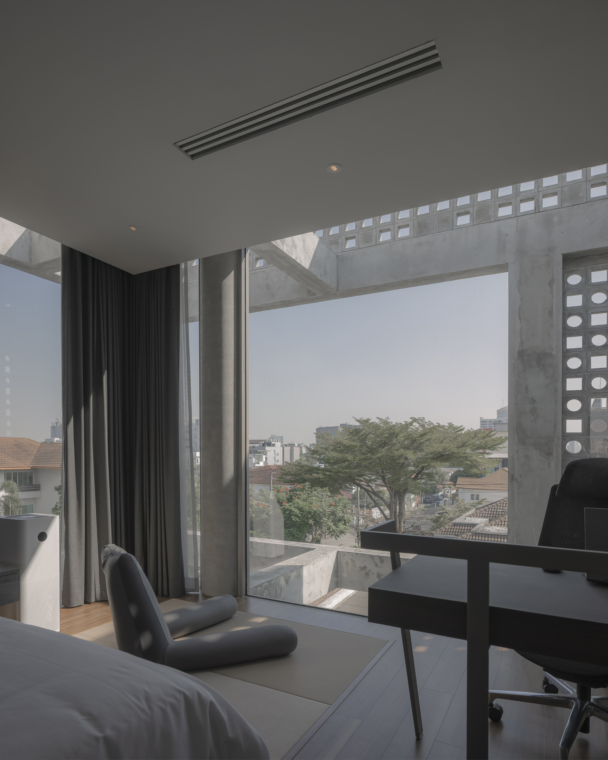 Bedroom window framed by perforated concrete beams overlooking neighborhood rooftops and a tree
