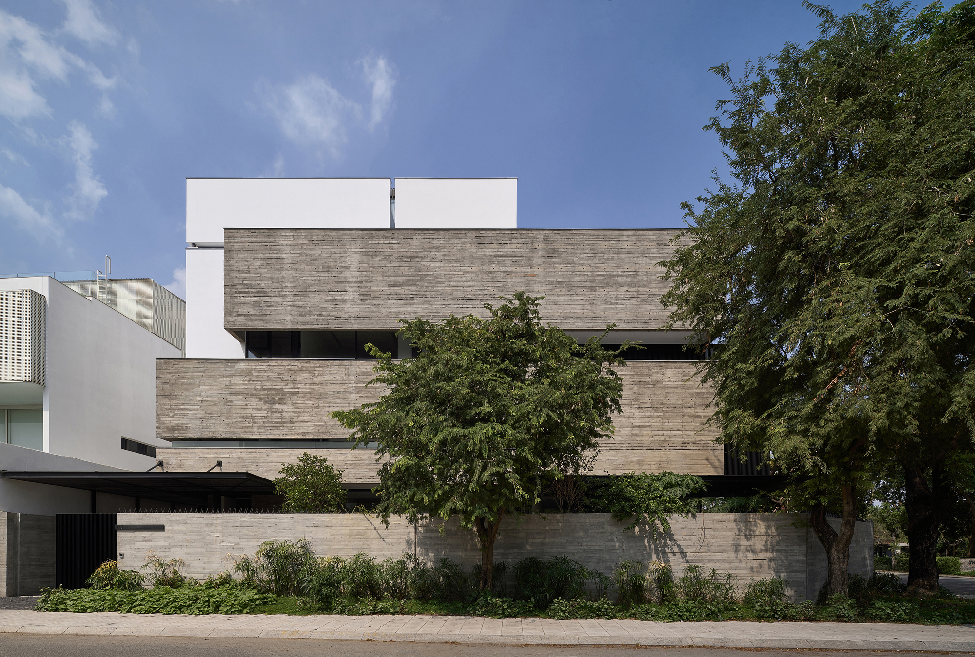 Street view of the stacked concrete facade with recessed windows behind planted beds and mature trees