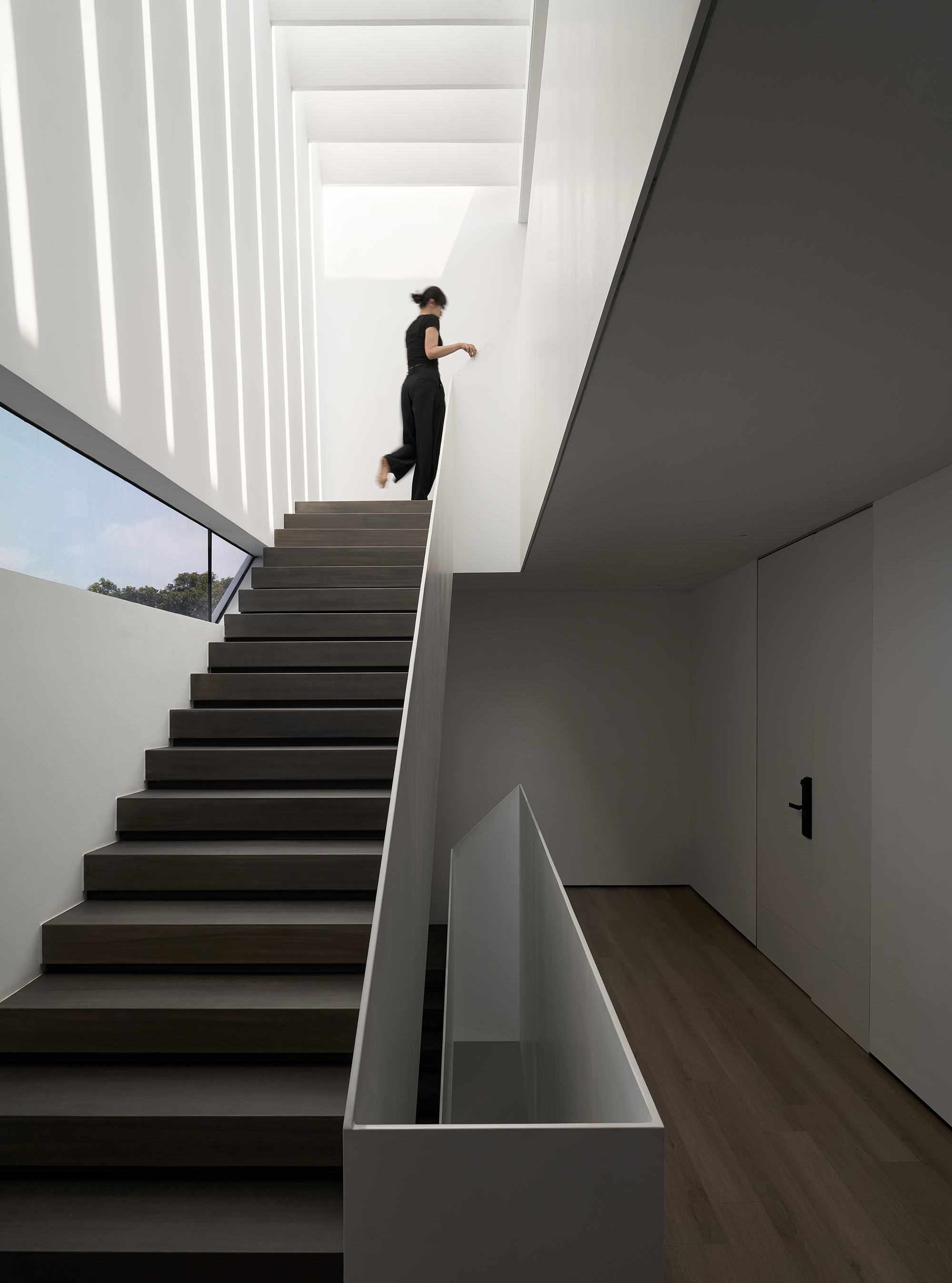Dark timber staircase with figure ascending under vertical skylights casting striped shadows