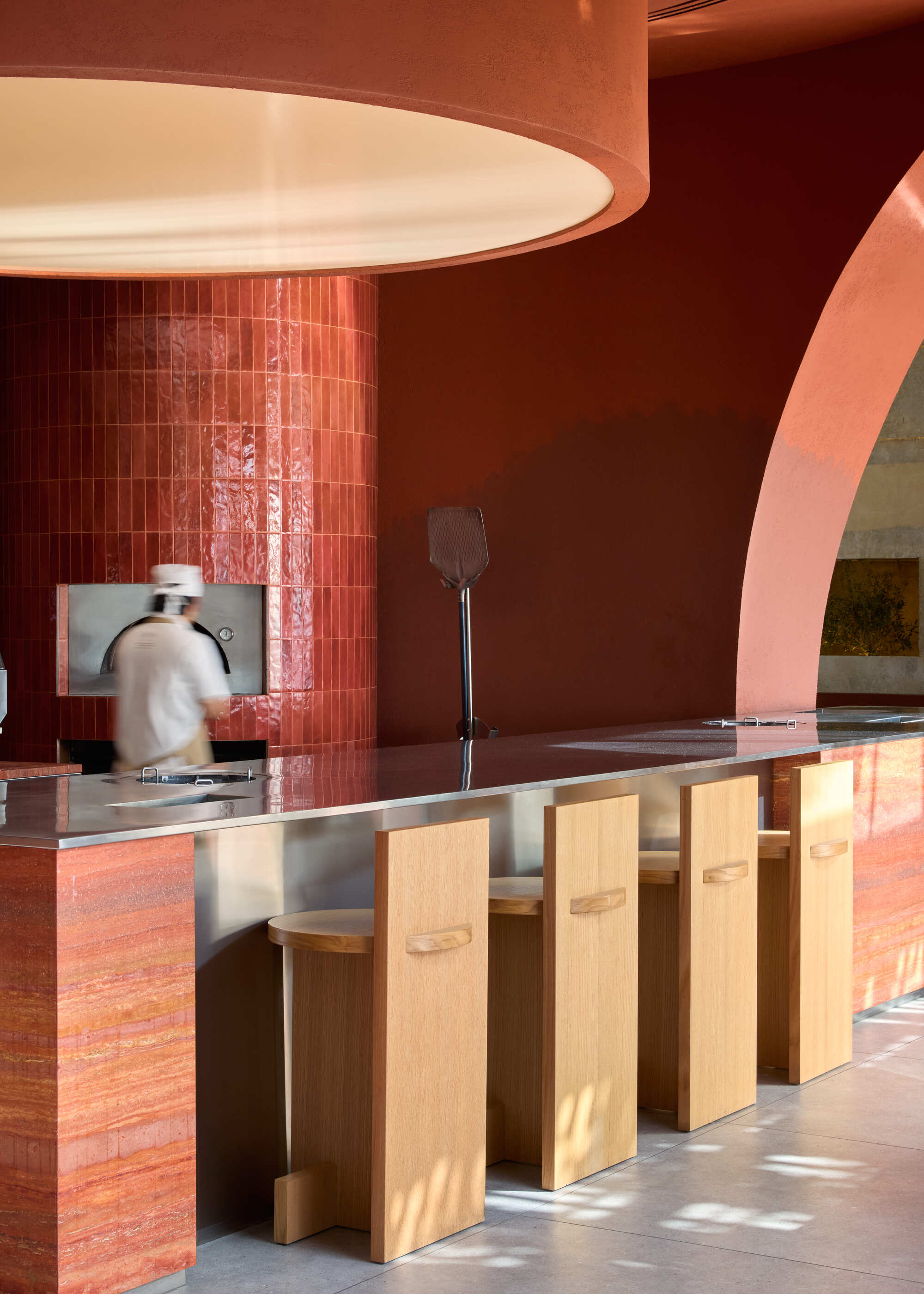 Counter with timber stools beneath a circular pendant light in a terracotta-toned interior with sunlight streaming in
