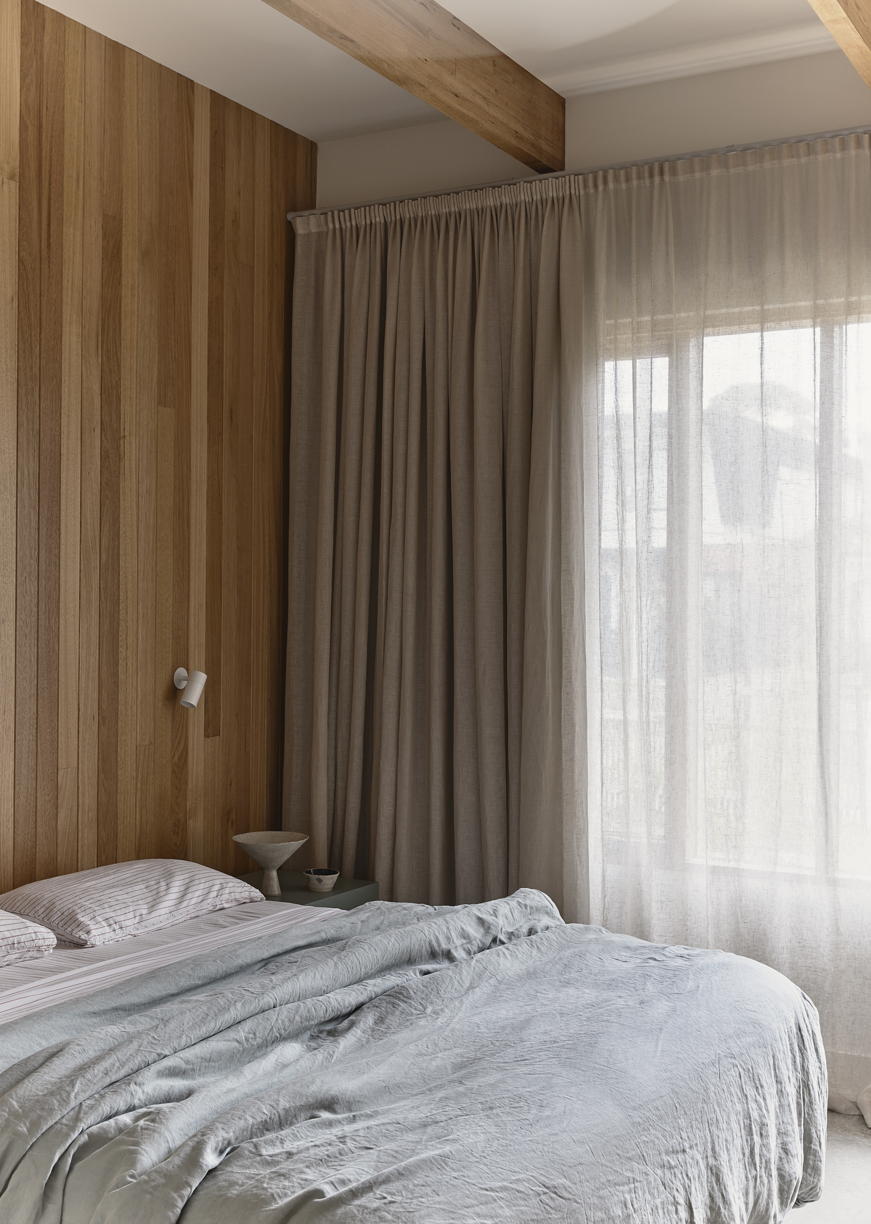 Bedroom with timber paneling and exposed beam ceiling beside layered linen curtains in soft light