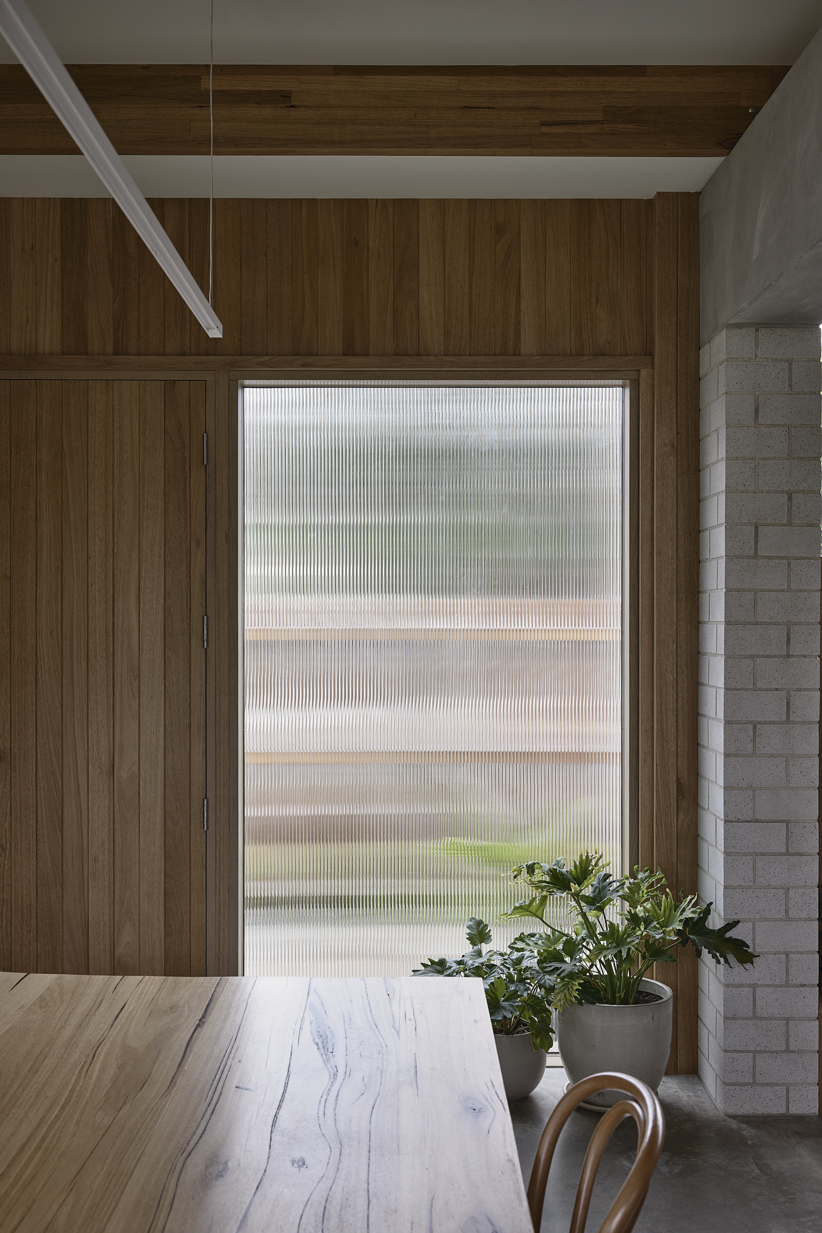 Dining table beneath fluted glass window filtering daylight alongside timber wall and concrete block edge
