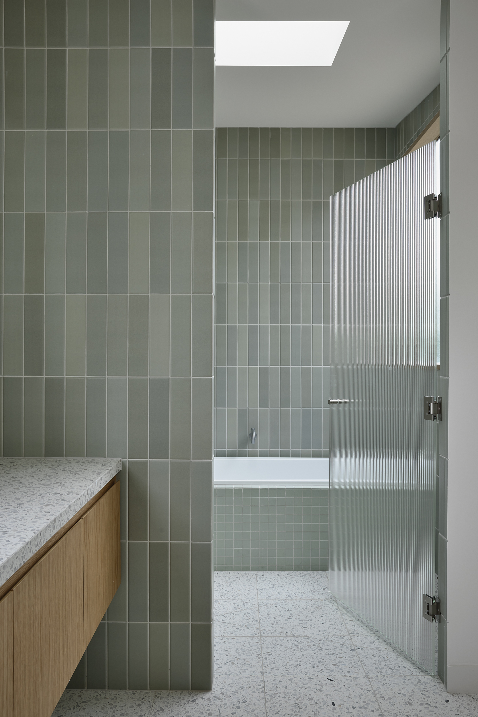 Bathroom interior with vertical sage green tile walls, fluted glass shower door, and terrazzo flooring under a skylight
