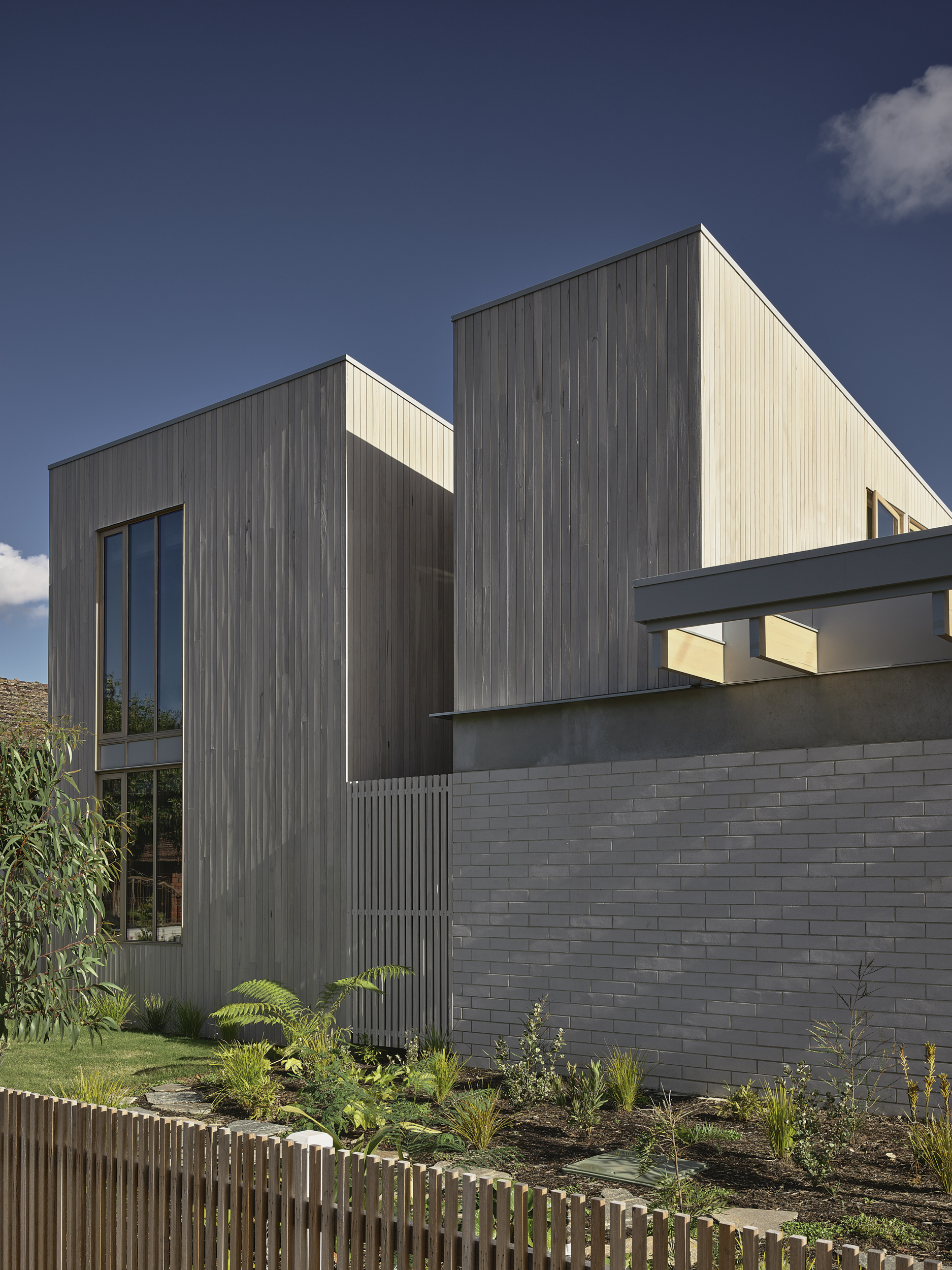 Street facade showing vertical timber cladding above a grey brick base with planted garden bed and timber fence