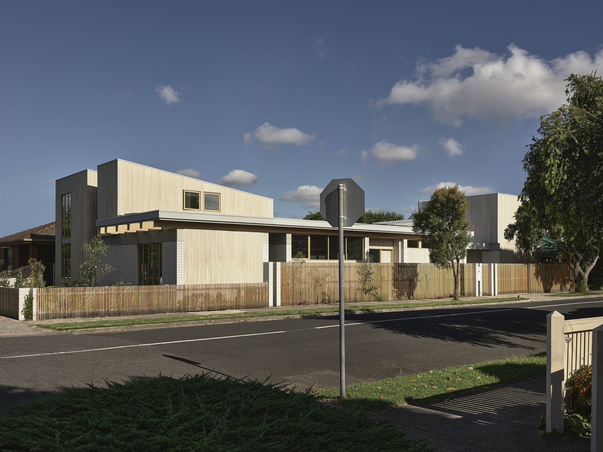 Street view of the residence with vertical timber cladding, timber fence, and mature street trees under scattered clouds