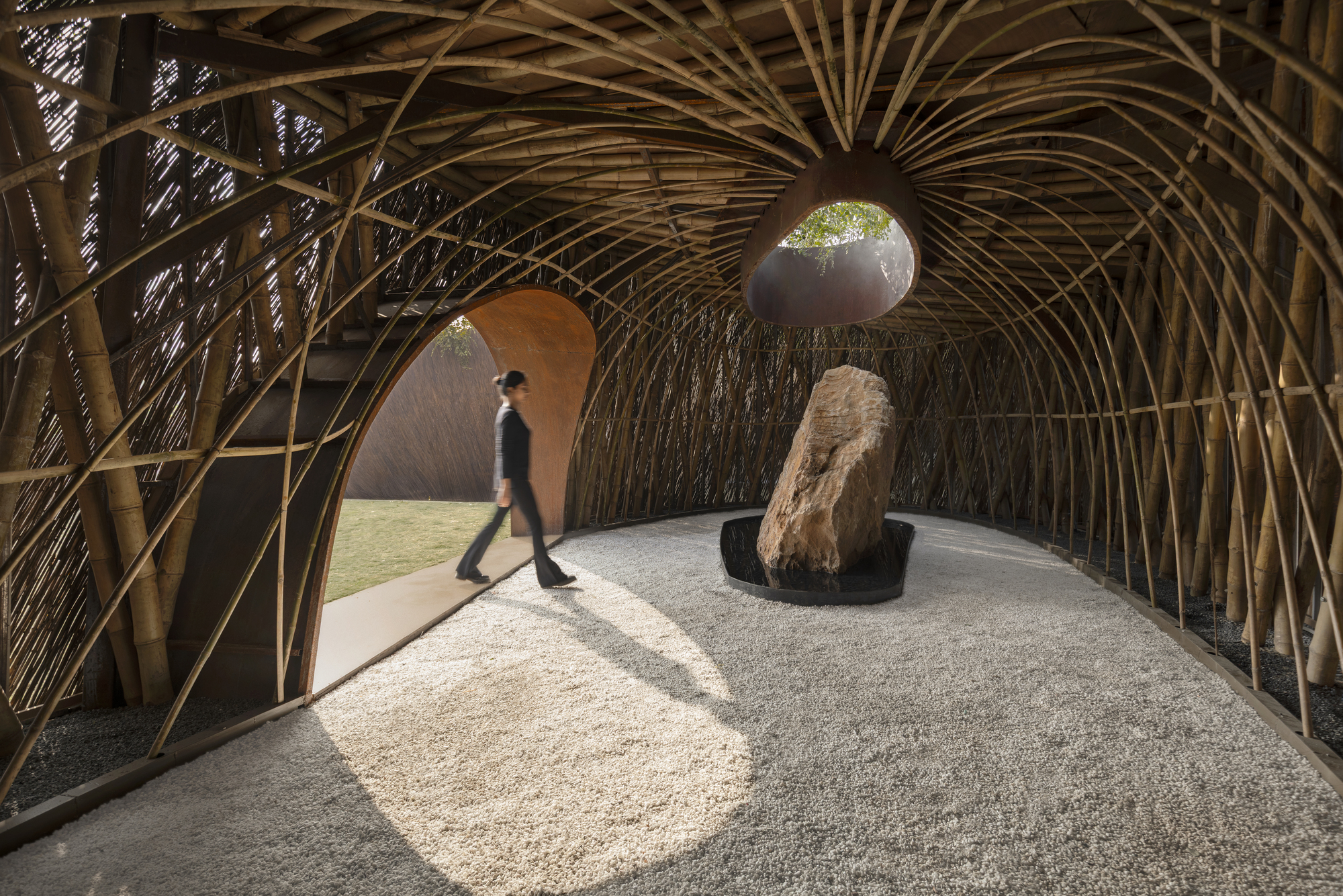 Vaulted bamboo interior structure with figure walking past boulder and gravel floor in dappled sunlight