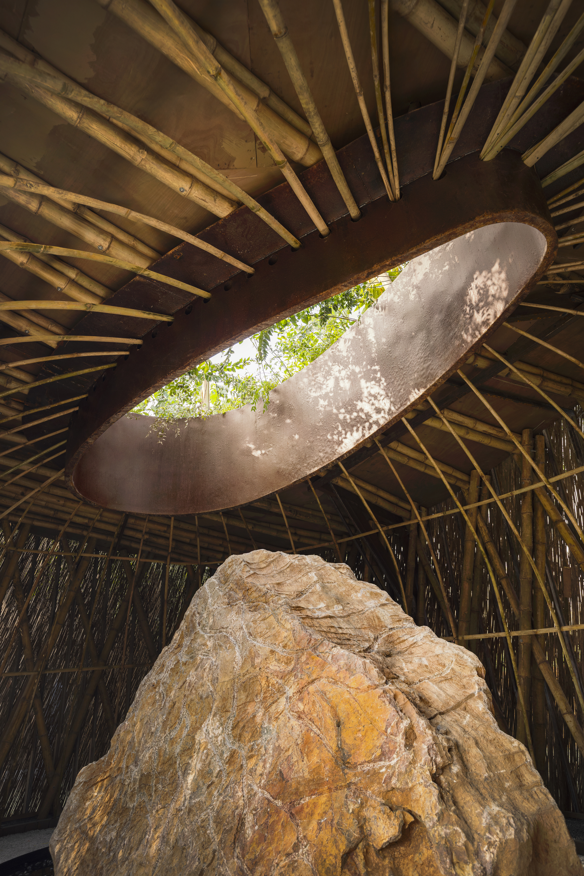 Looking up through oval roof opening with radiating bamboo structure and weathered boulder catching dappled sunlight below