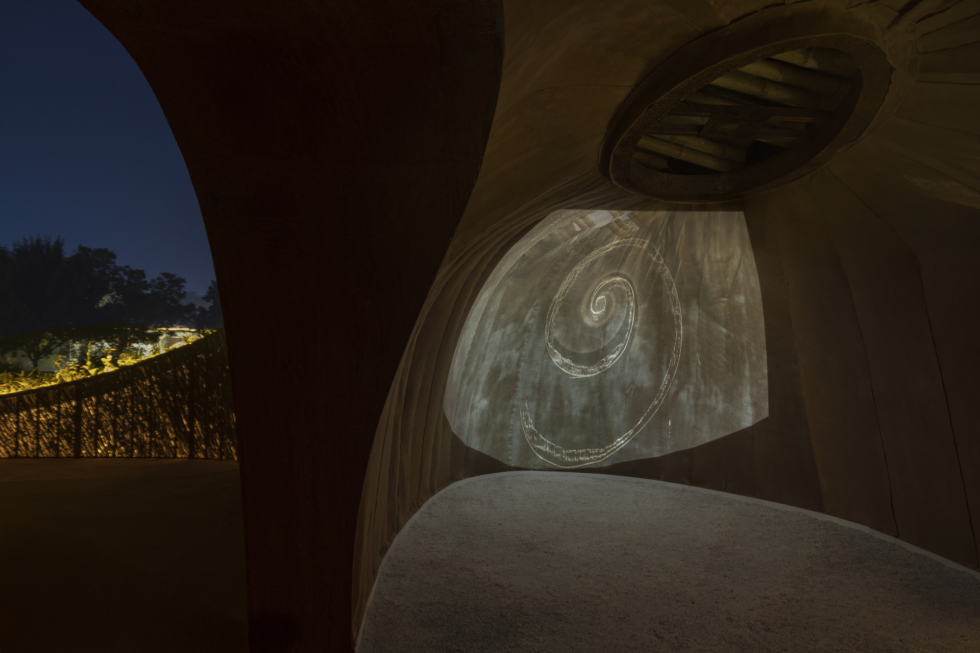 Curved illuminated wall with spiral relief pattern between arched openings and oval roof apertures at dusk