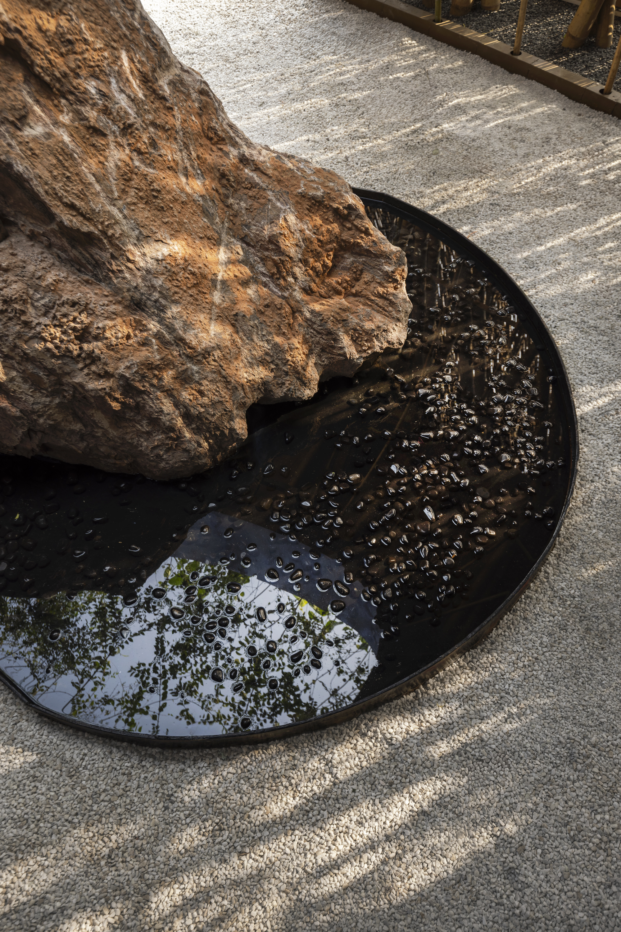 Close-up of weathered boulder resting on circular black reflecting pool with gravel floor casting rippled reflections