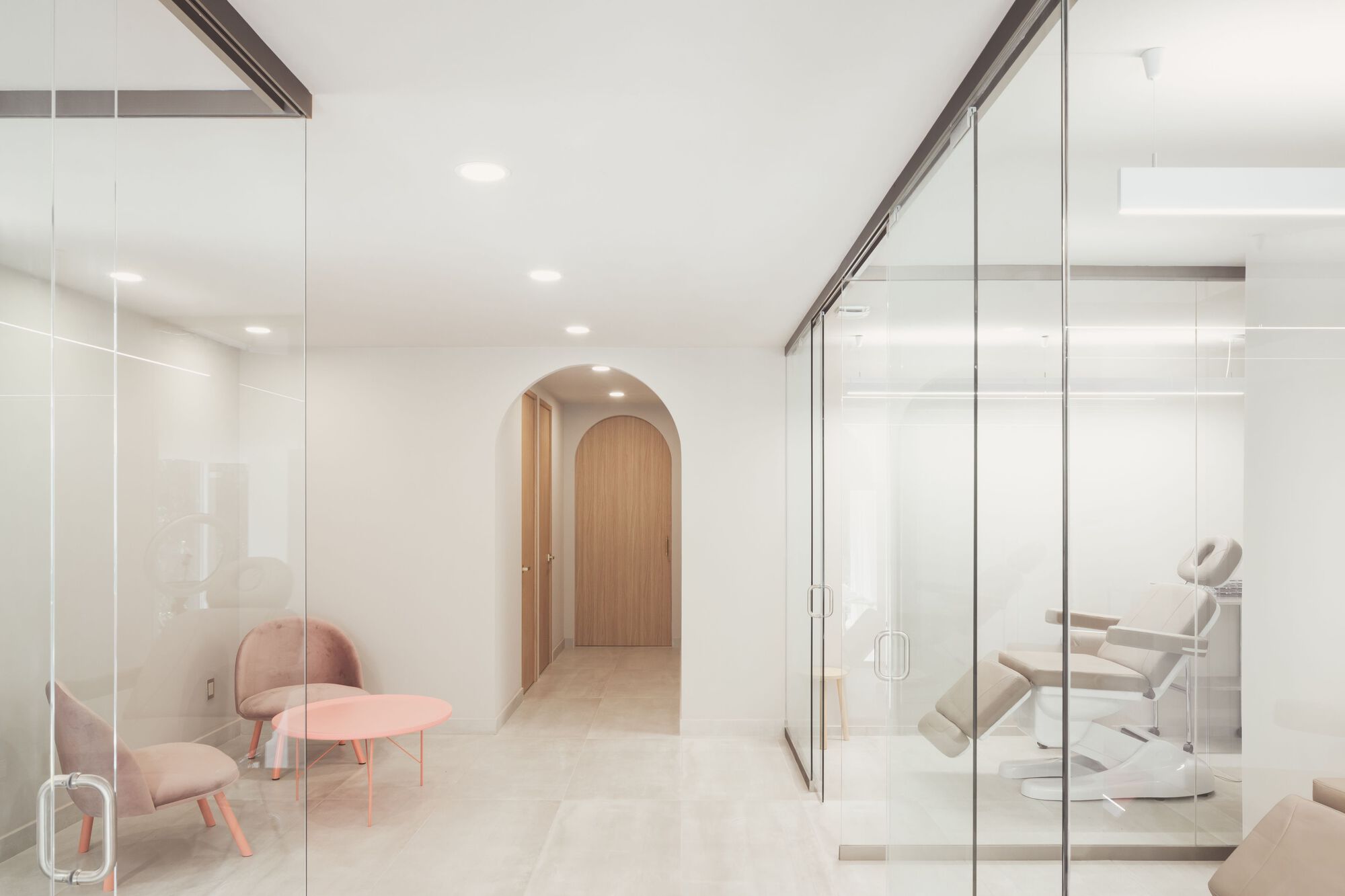 Corridor with floor-to-ceiling glass partitions revealing treatment rooms and a timber-clad arched doorway beyond
