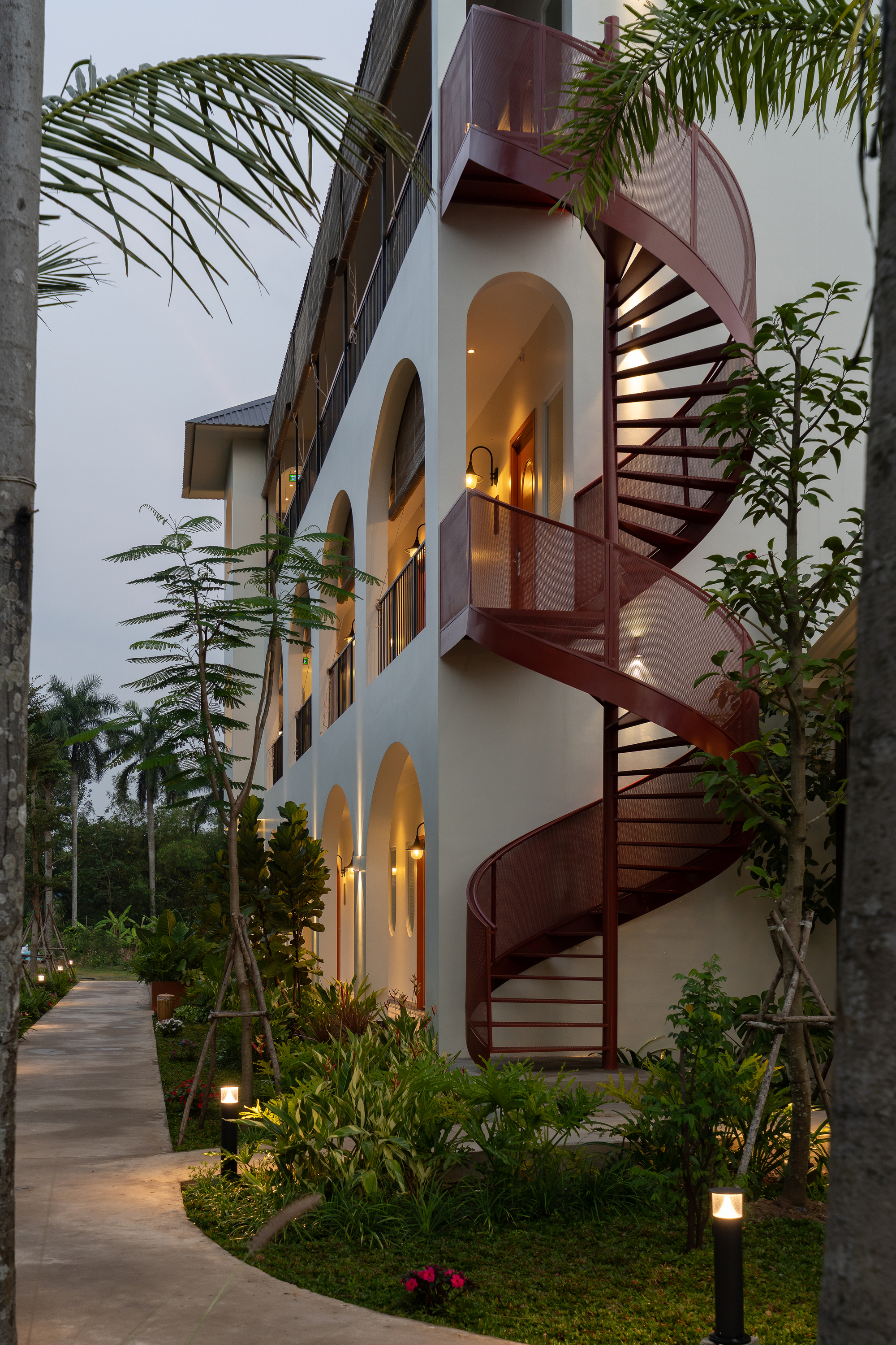 Exterior red steel spiral staircase ascending three stories along the white stucco facade at twilight