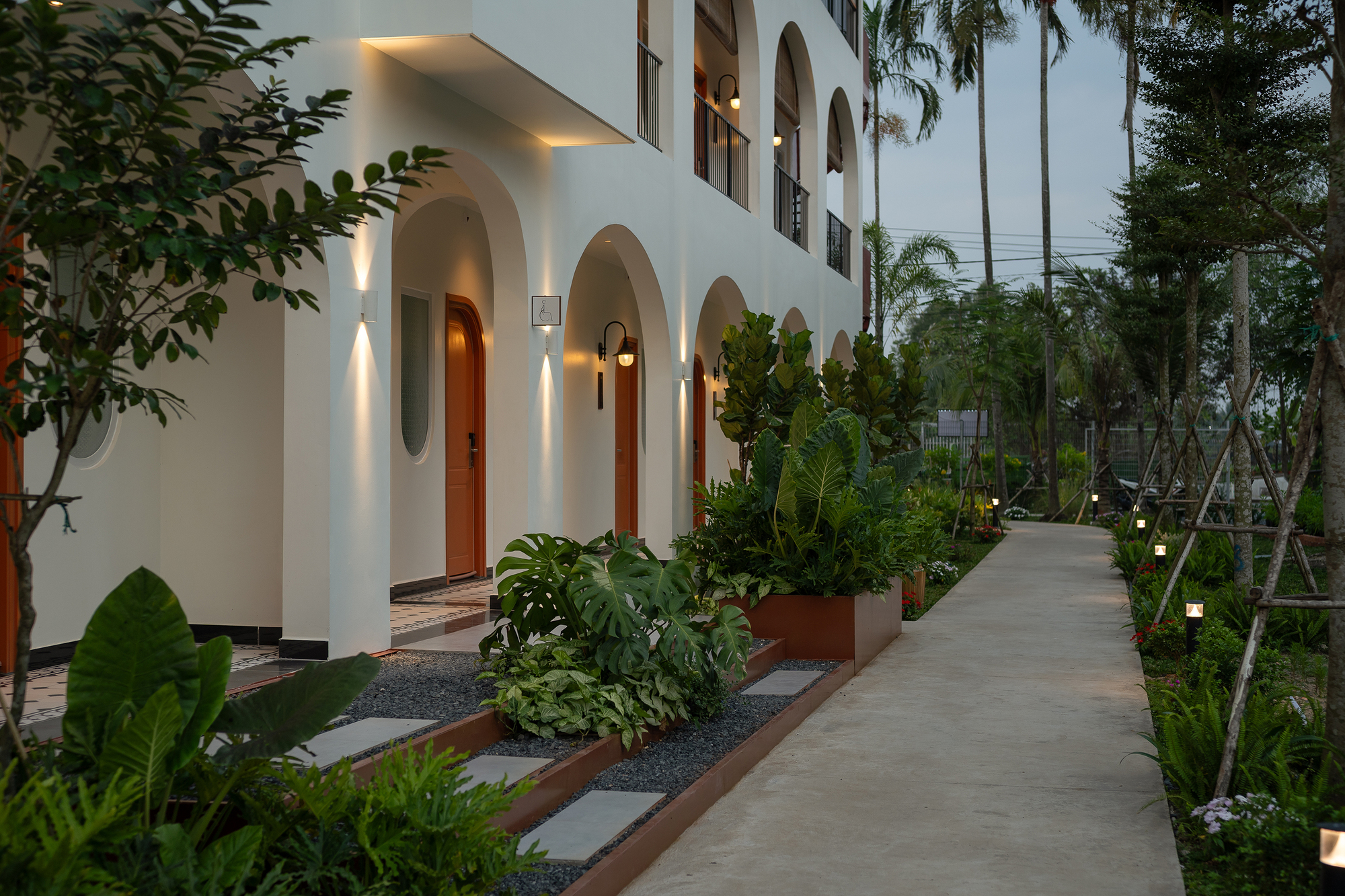 White arched facade with planted beds and concrete walkway at dusk under palm canopy