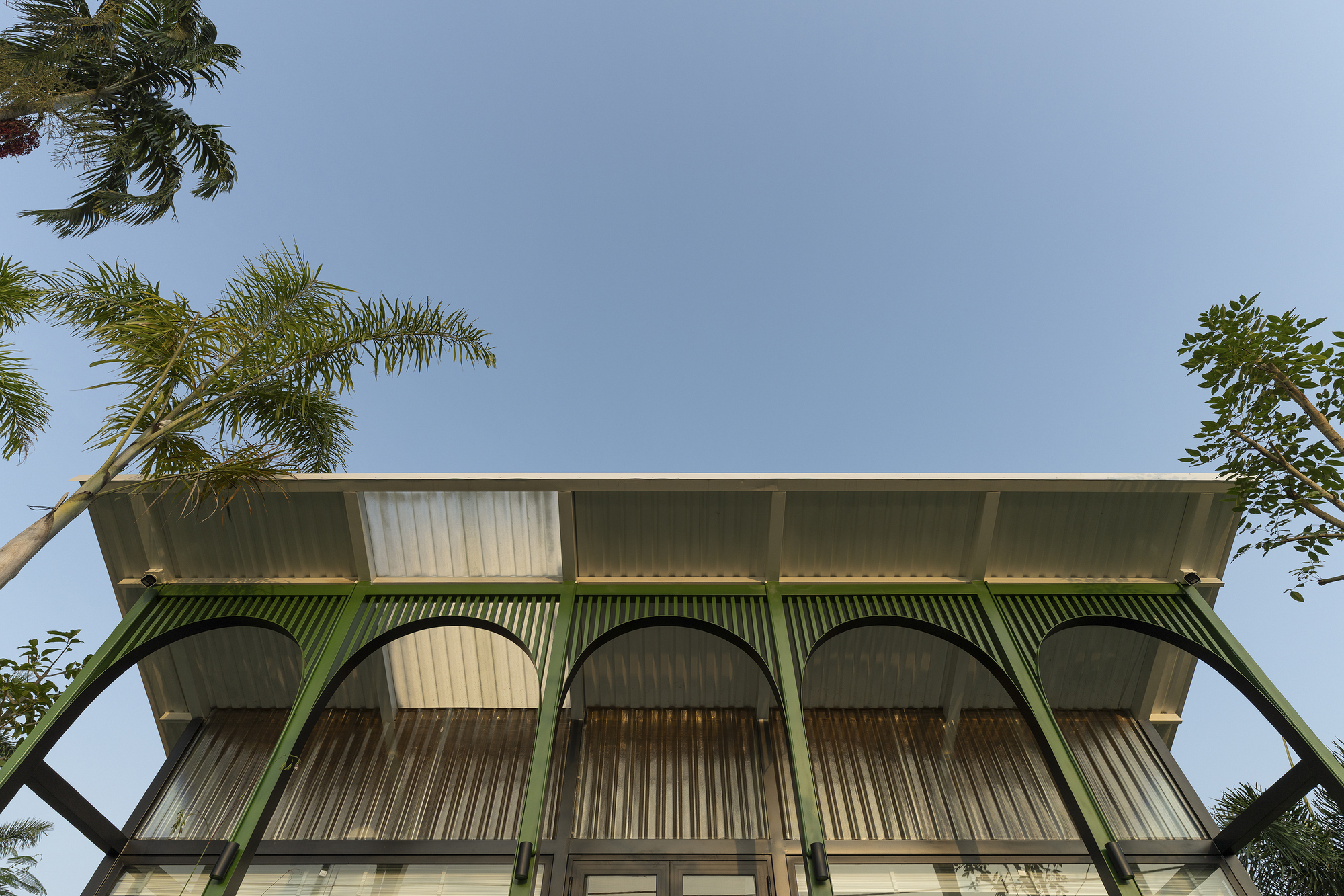 Upward view of the corrugated metal facade with green arched supports framed by palm fronds