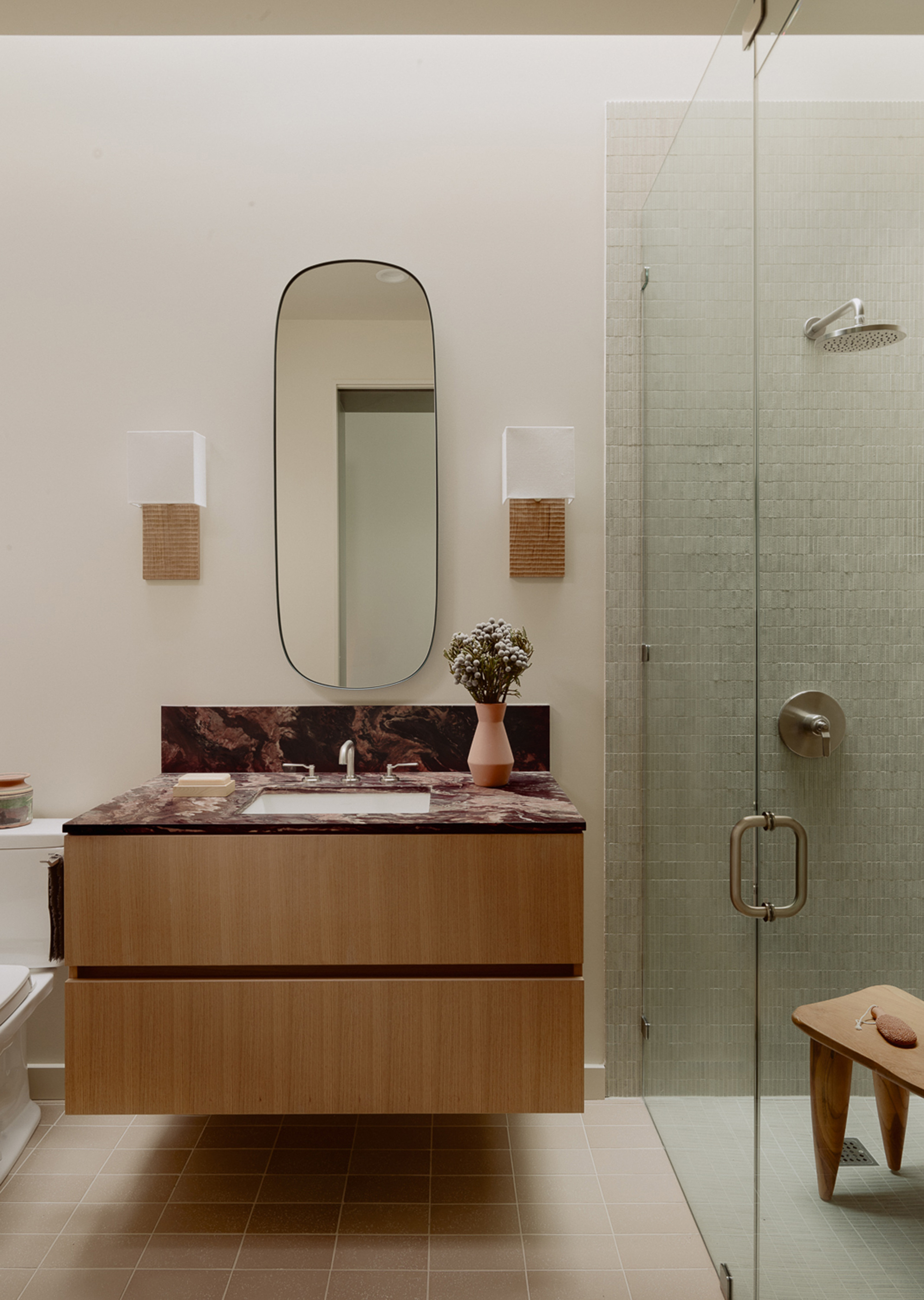 Floating oak vanity with marbled countertop beside a glass-enclosed shower with pale green mosaic tile