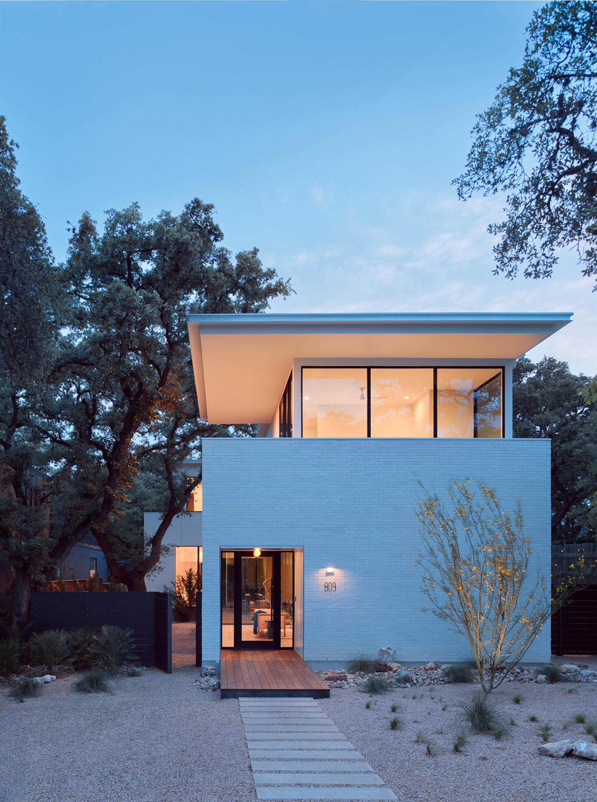 Two-story white brick volume with floor-to-ceiling glass doors illuminated at dusk beside an old oak tree