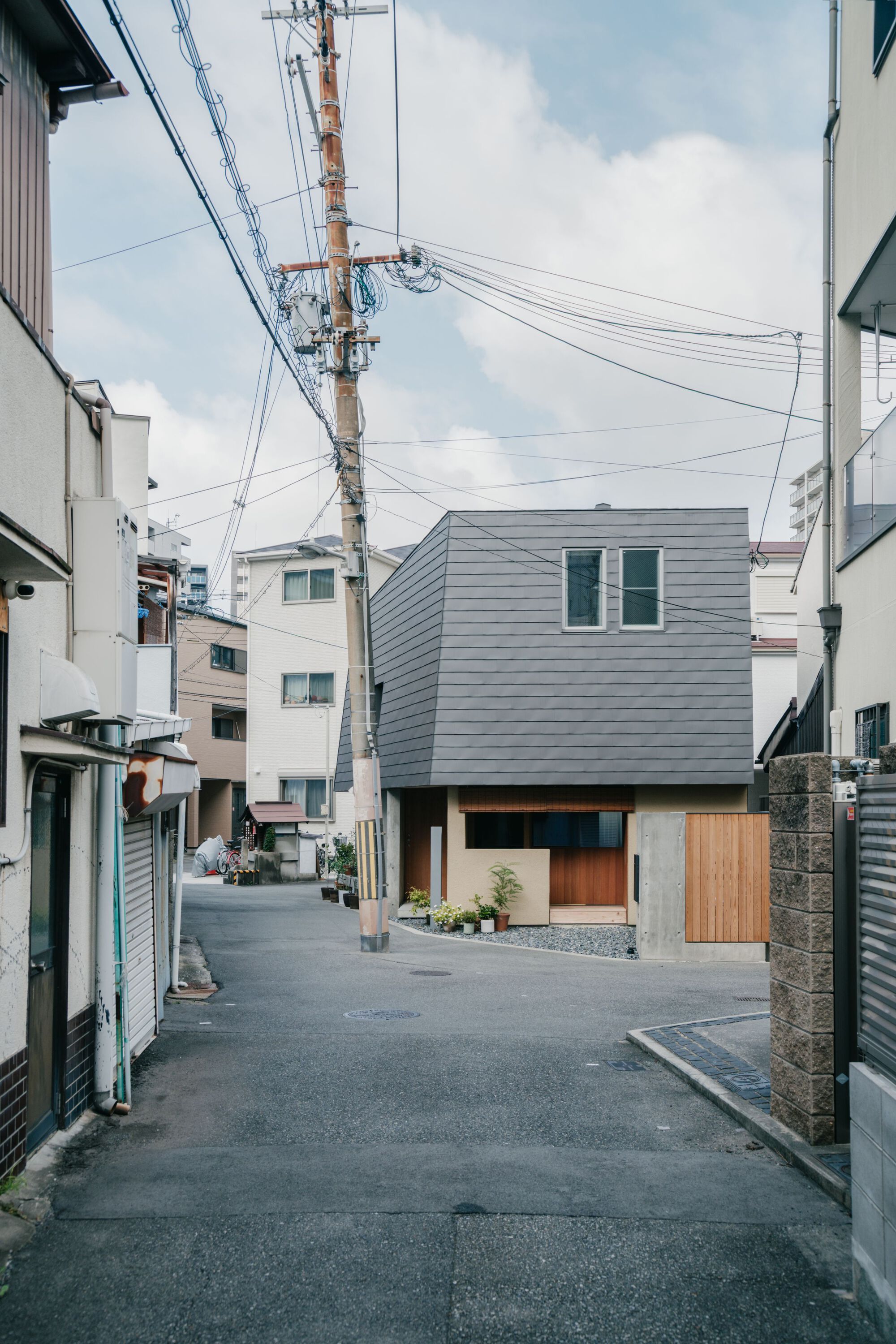 Neighborhood street view showing the cantilevered upper volume clad in grey metal panels above timber and plaster base