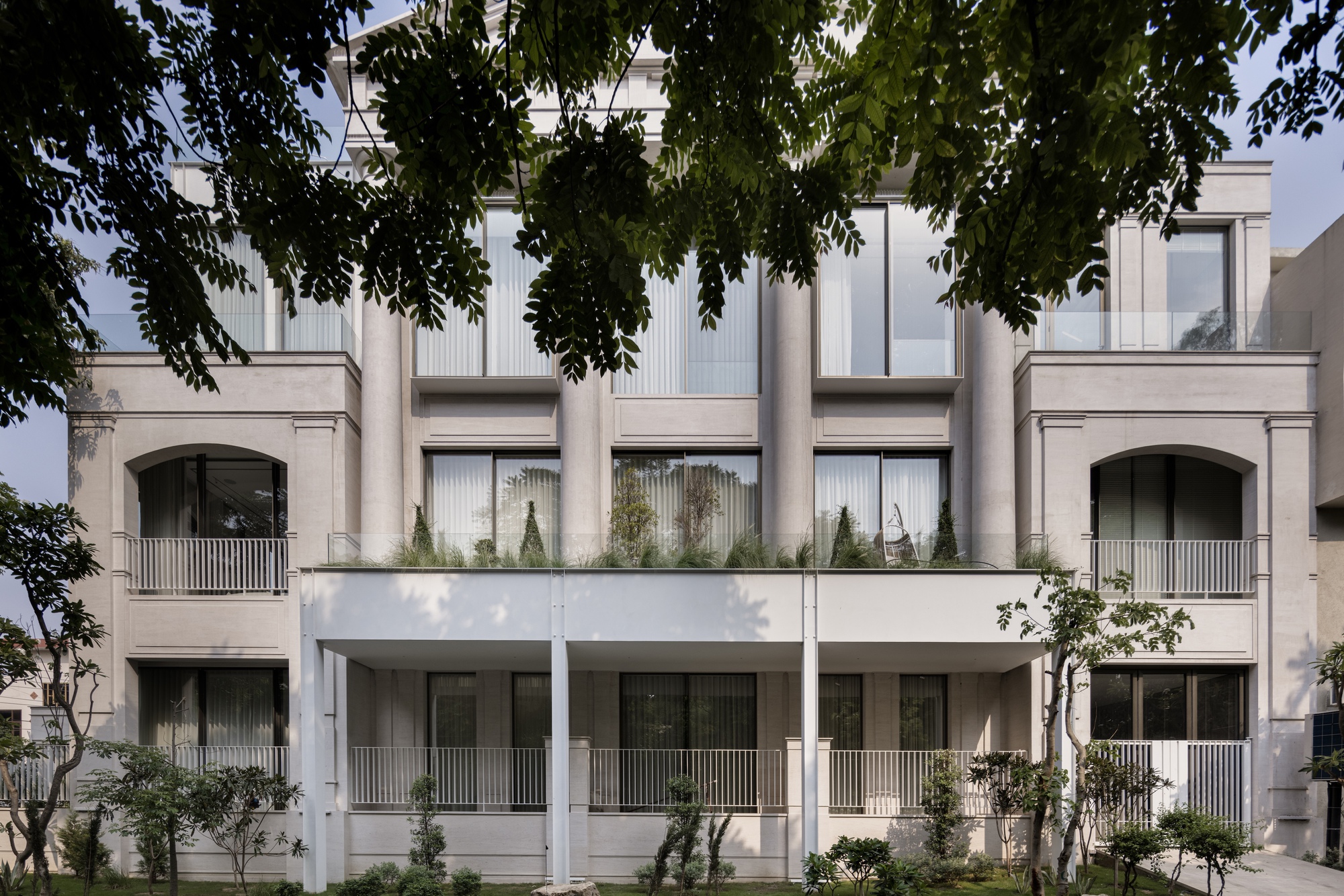 Street facade with white balconies, planted terraces and arched openings framed by overhanging tree branches