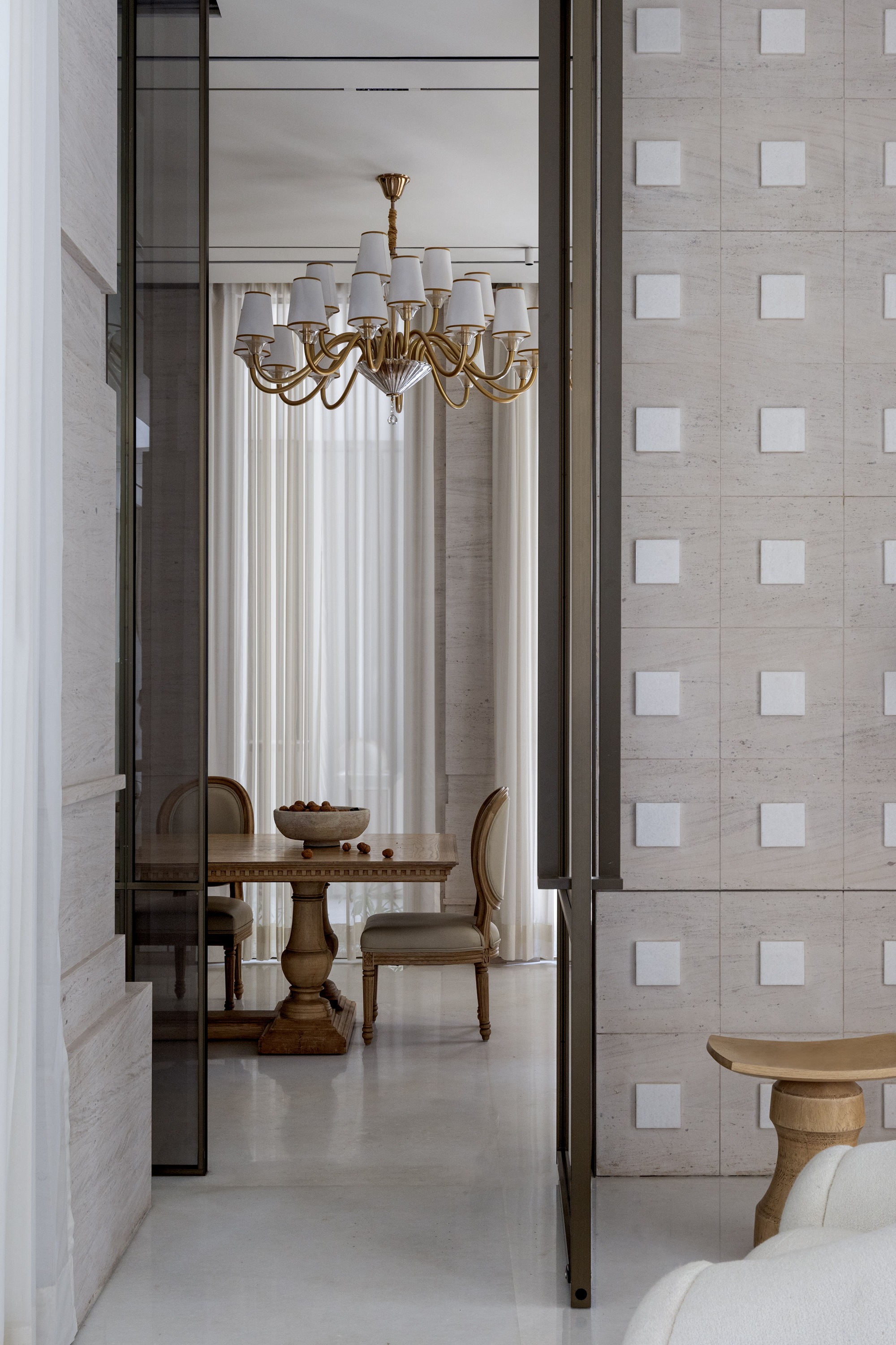 View through bronze-framed glass doors to dining room with brass chandelier and stone grid wall