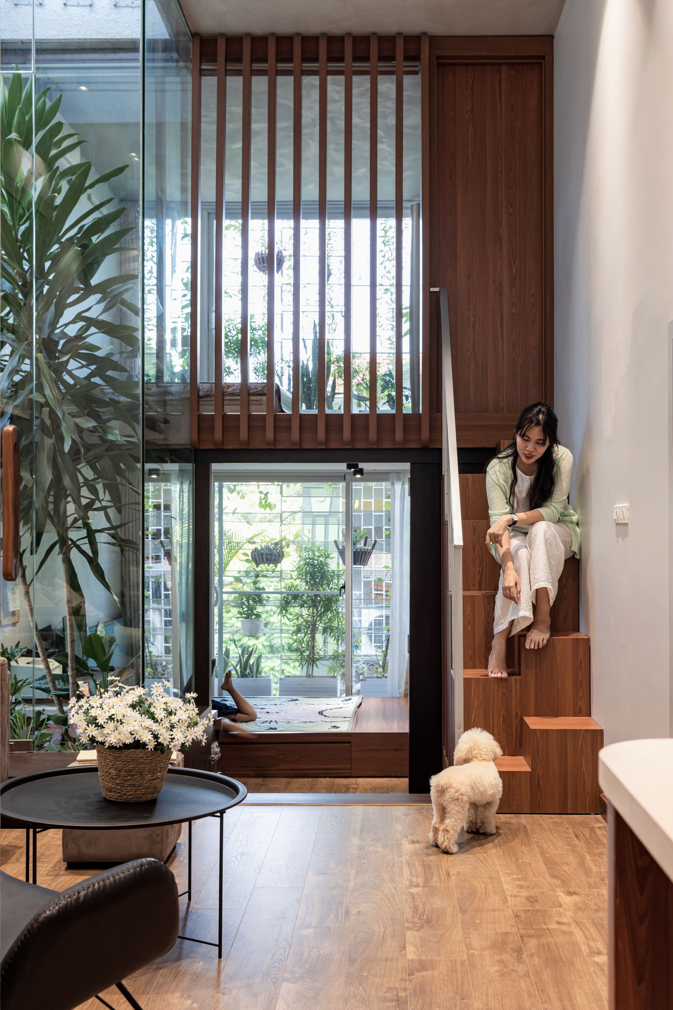 Double-height living room with timber slatted screen and woman seated on wooden stair beside dog