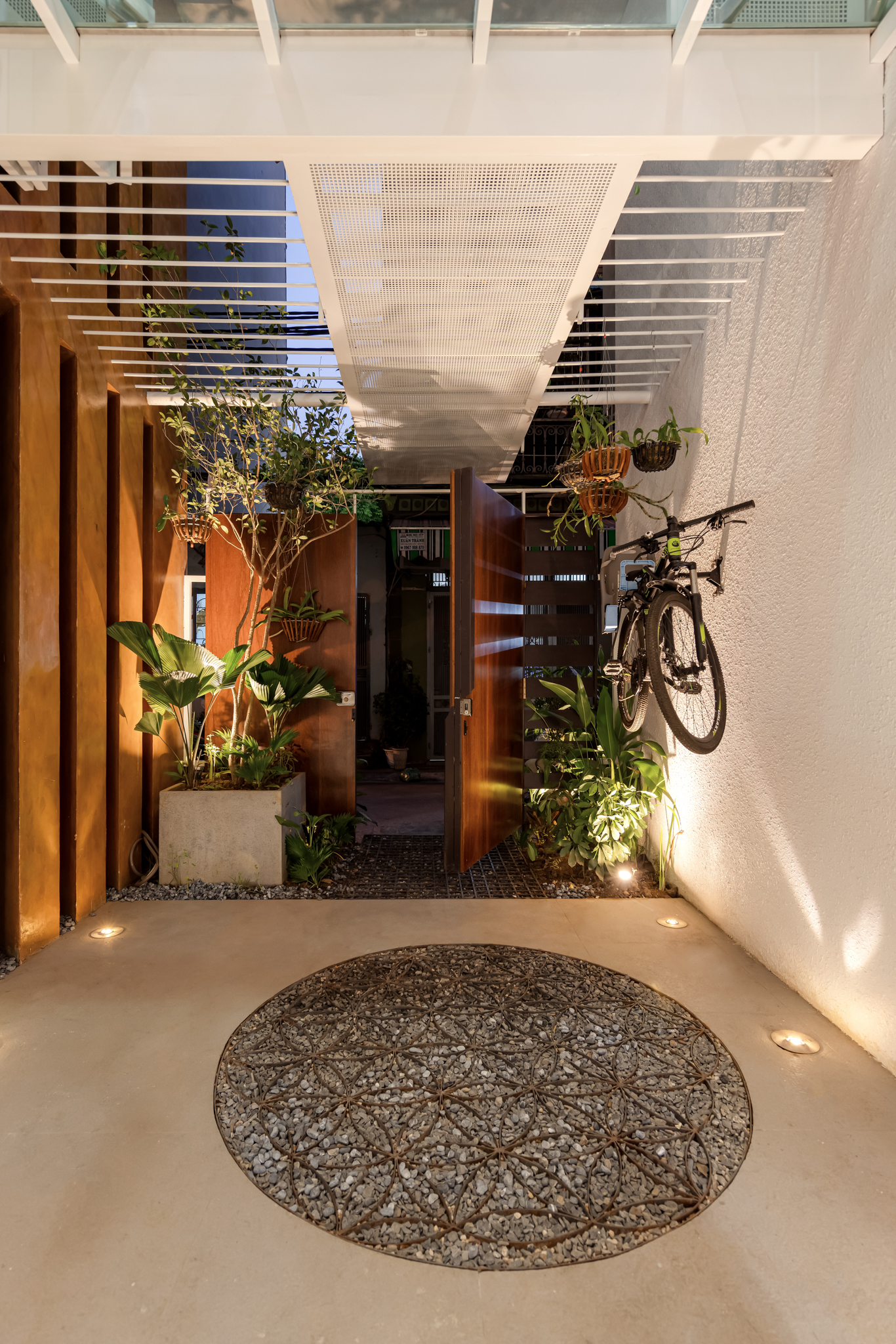 Entry courtyard at dusk with circular pebble feature, timber screens, and potted plants under perforated ceiling