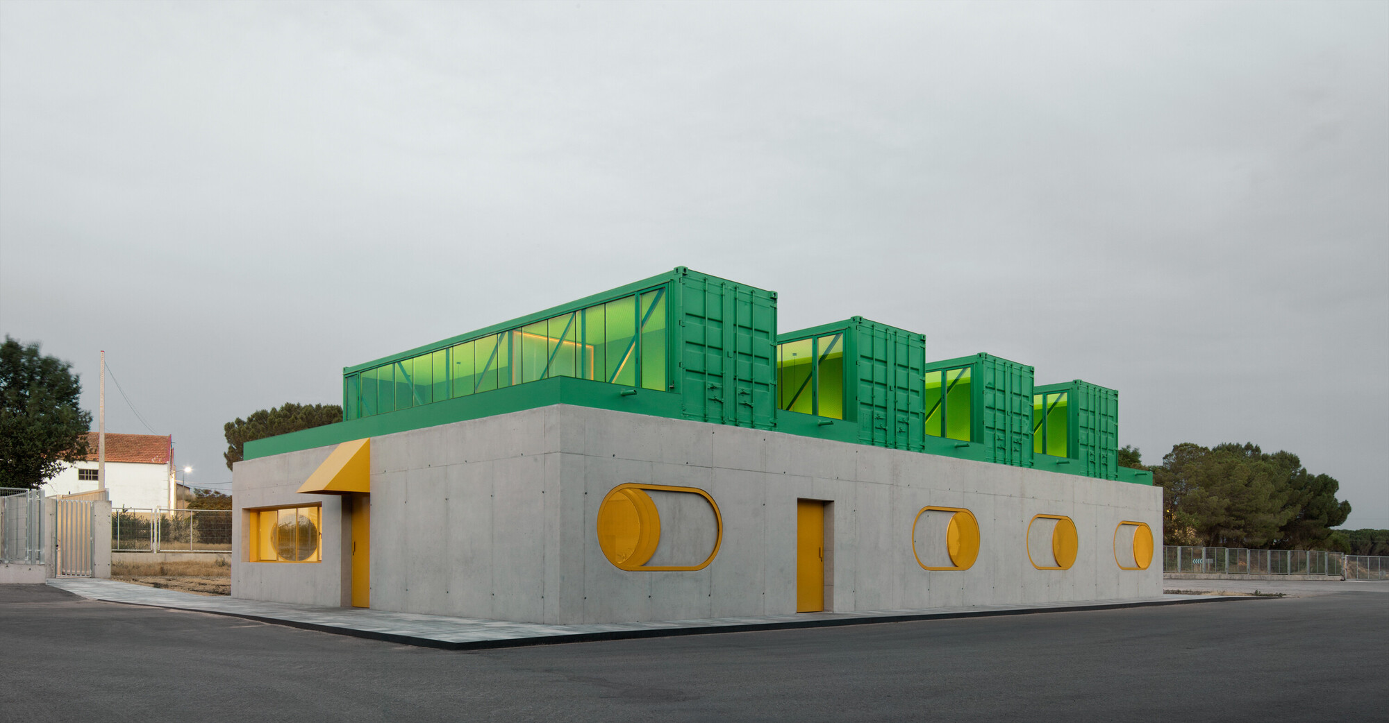 Corner view of concrete volume with circular yellow windows and green shipping containers under grey sky