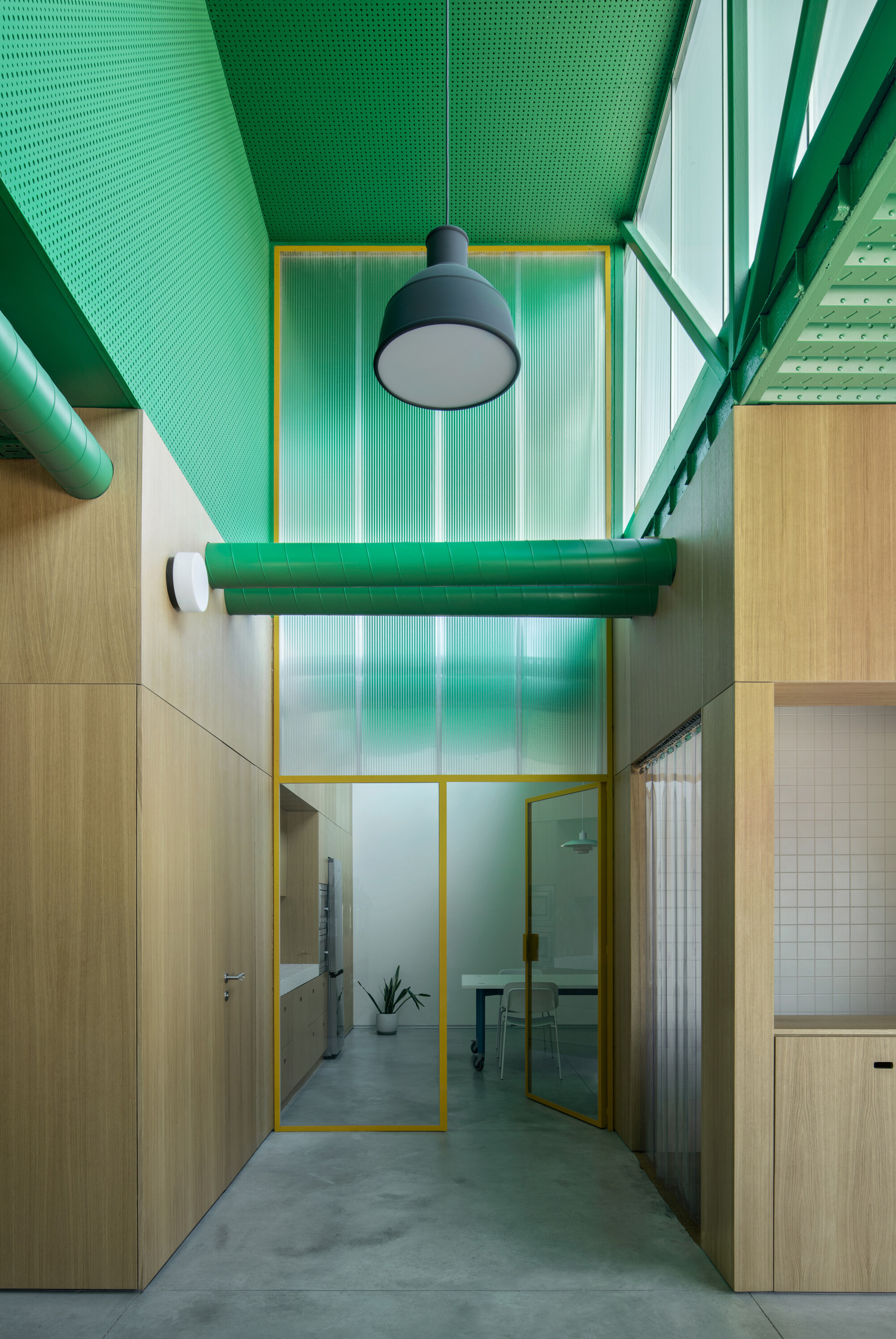 Double-height corridor with translucent green polycarbonate wall, yellow steel frame doorway, and exposed ductwork overhead