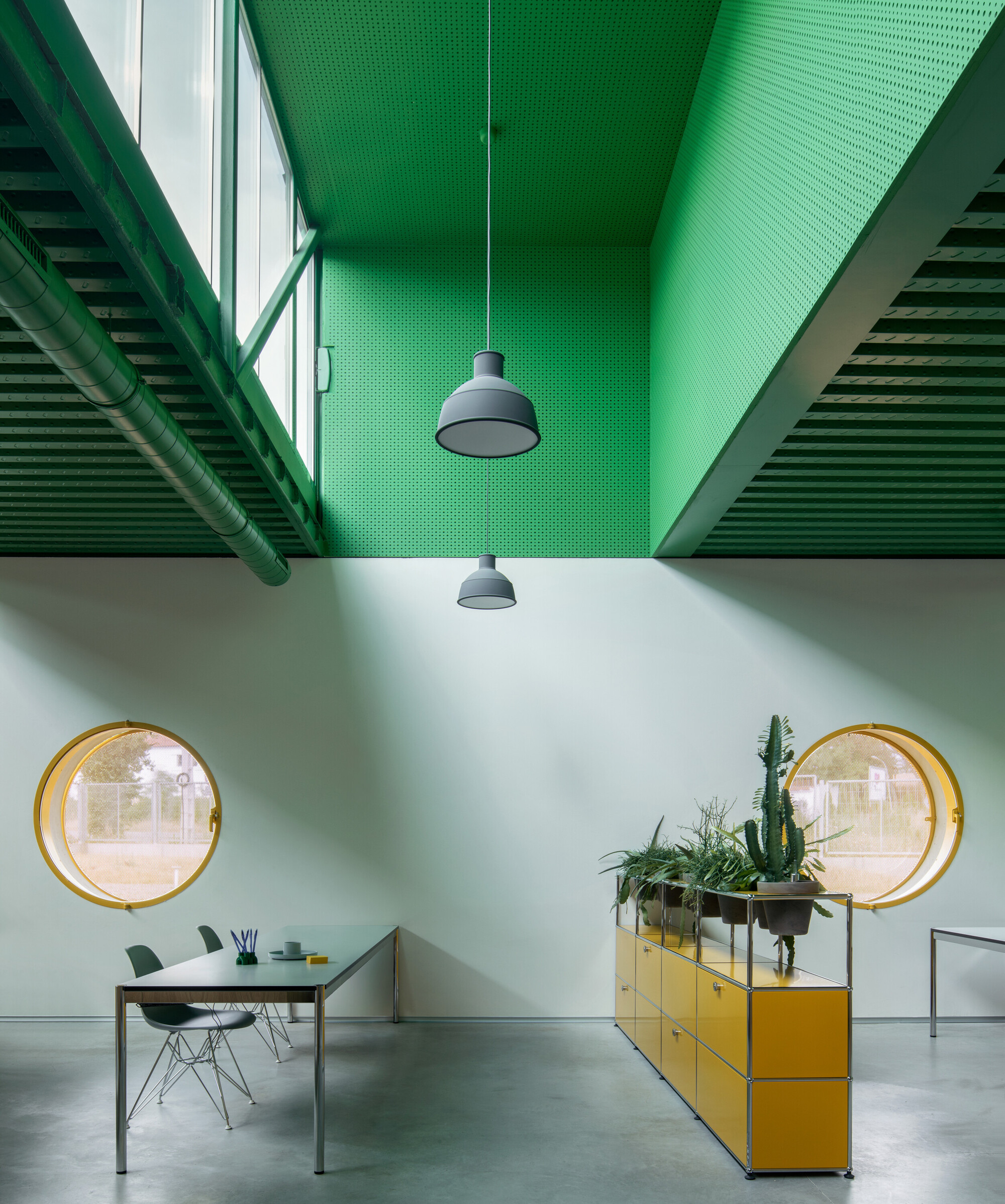 Double-height room with paired circular windows, green perforated ceiling, and potted cactus on yellow storage unit