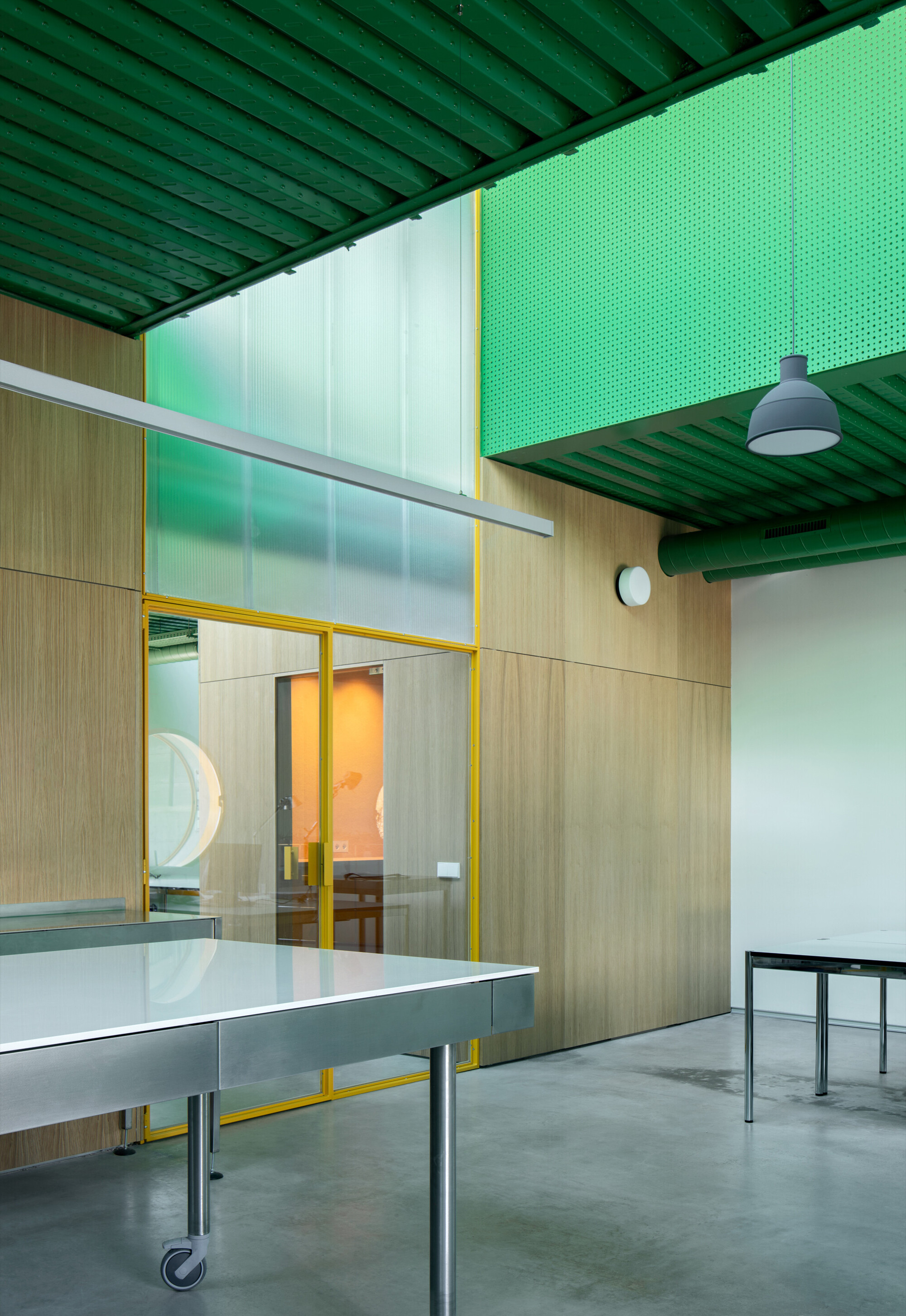 Interior workspace with yellow-framed glass partition, wood-clad walls, exposed green ceiling joists, and wheeled white table