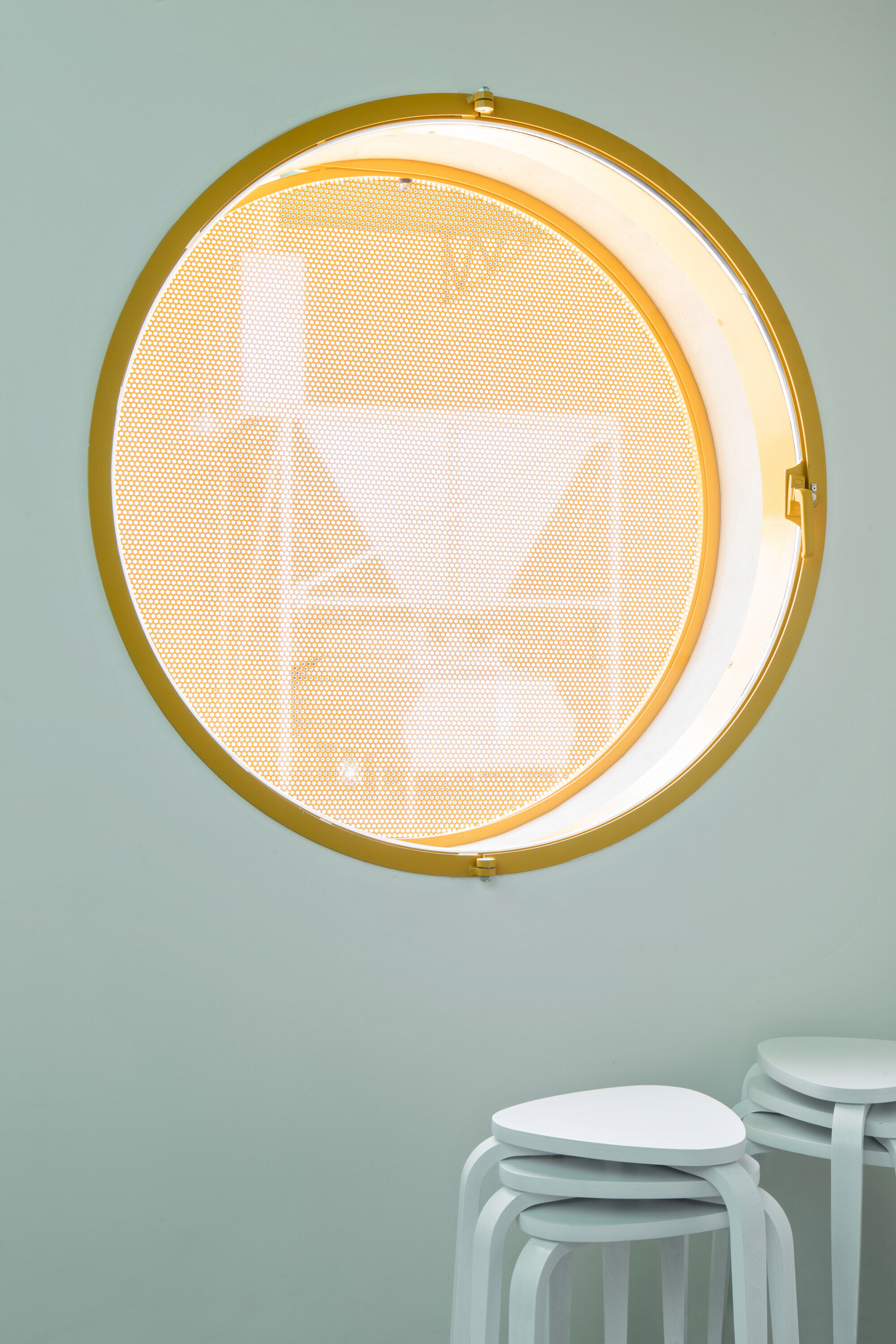Circular window with yellow metal frame and perforated screen on pale green interior wall