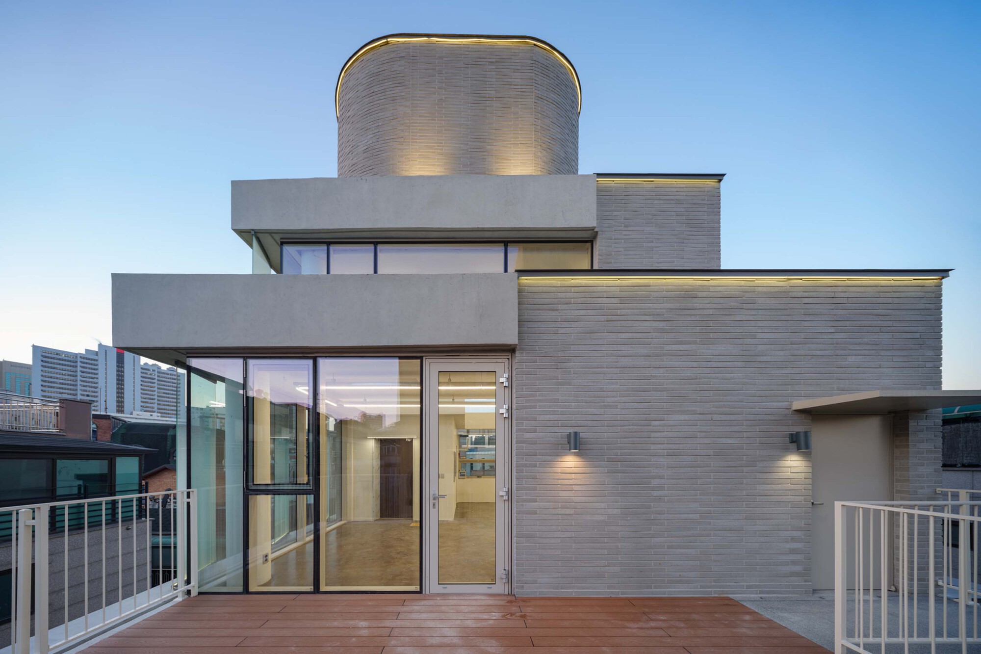 Rooftop terrace with timber decking, white railings, and curved grey brick tower illuminated at dusk