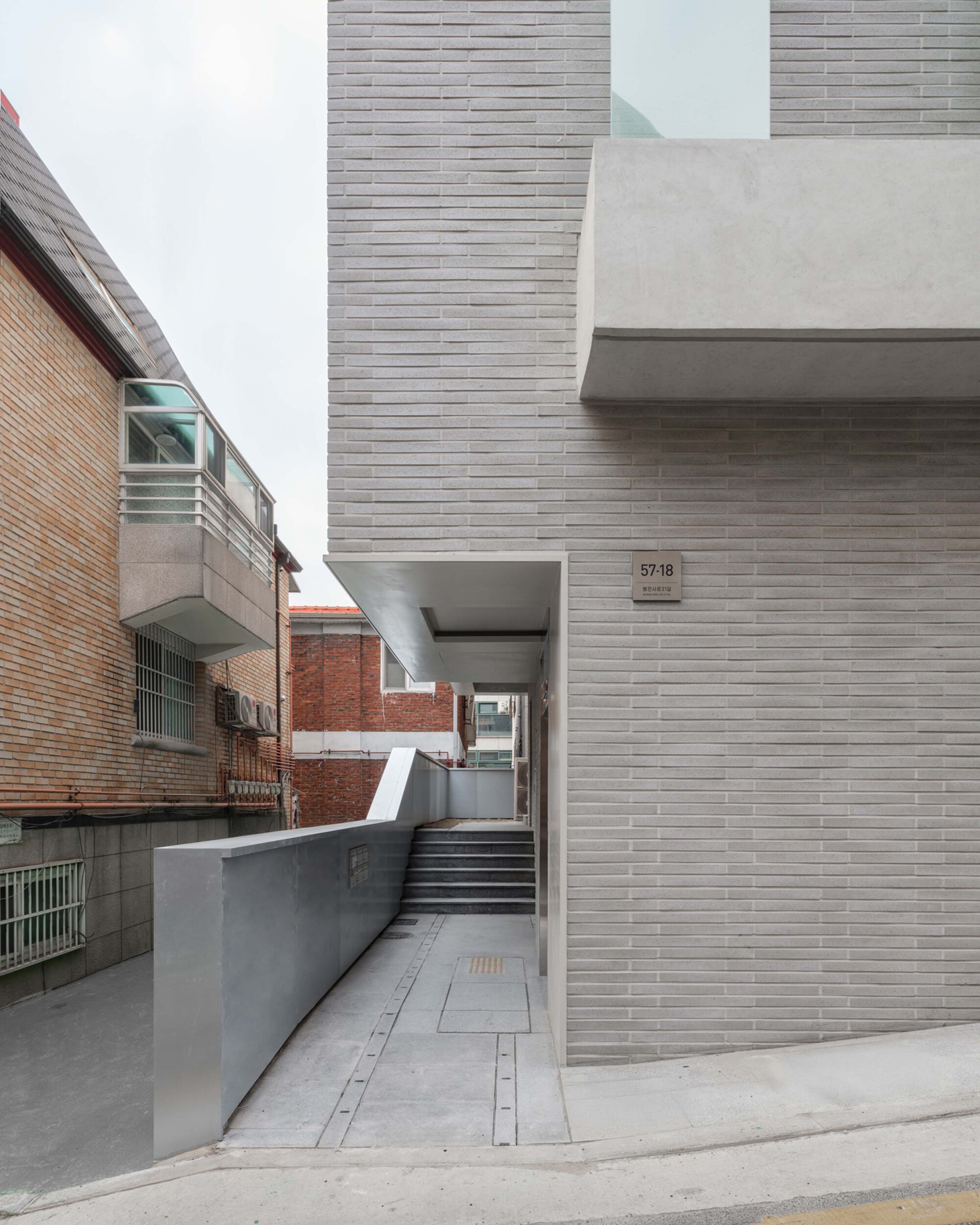 Pedestrian entry passage with horizontal grey brick cladding and a cantilevered concrete overhang
