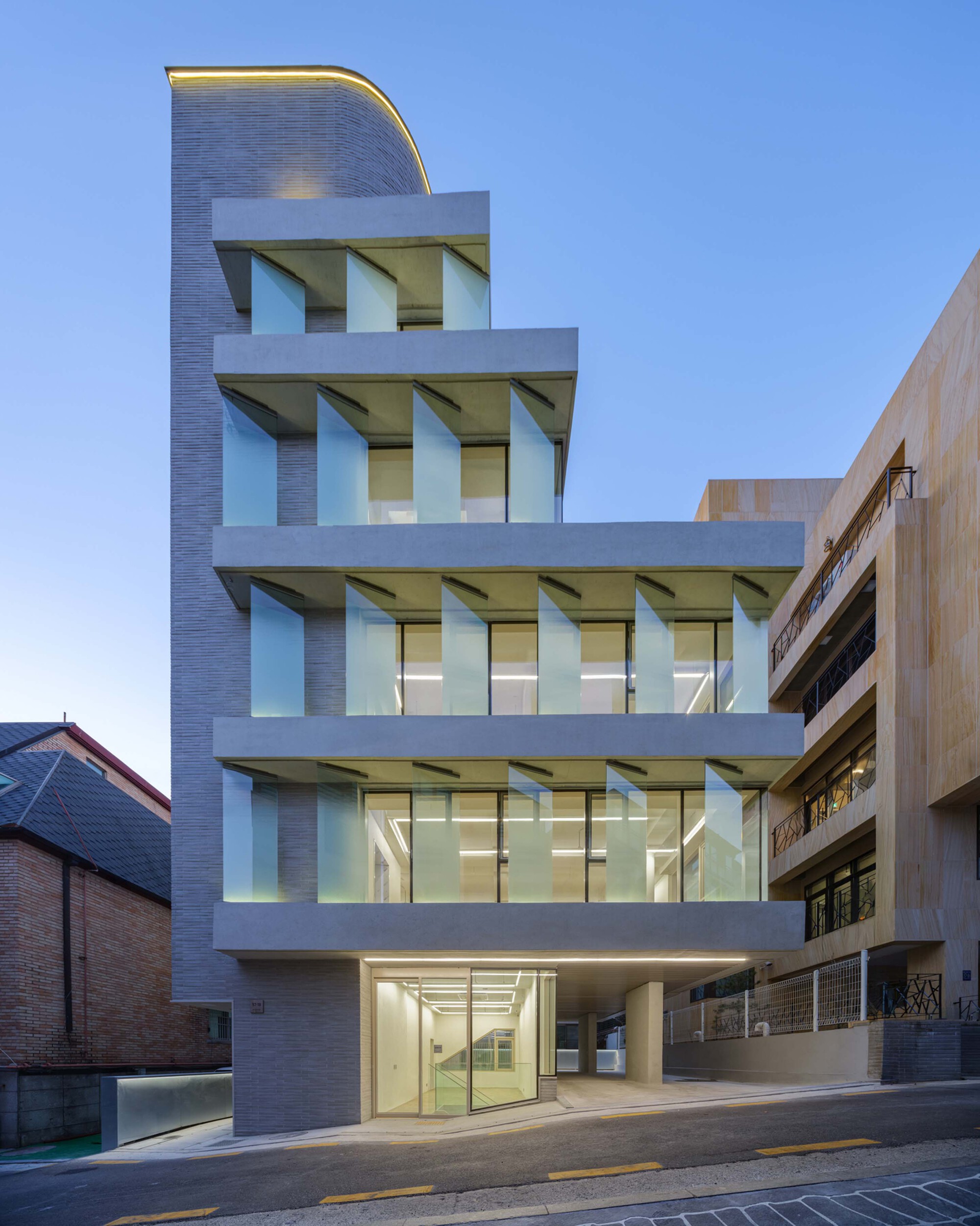 Street facade showing concrete slabs, angled sunshades and a curved brick tower at dusk