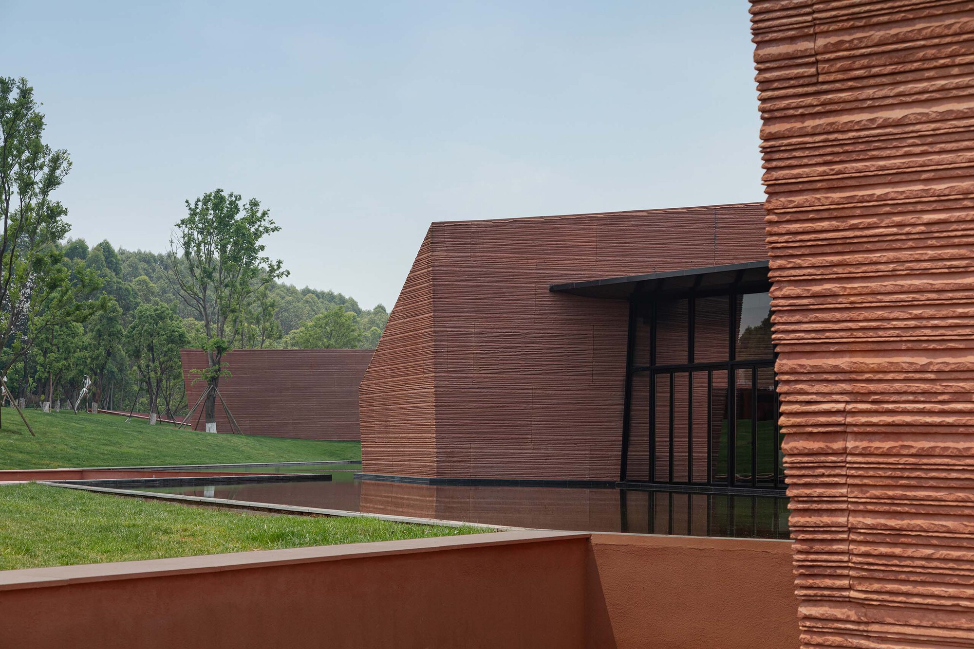 Horizontally striated red terracotta panels wrapping curved volumes with glass entry and reflecting pool
